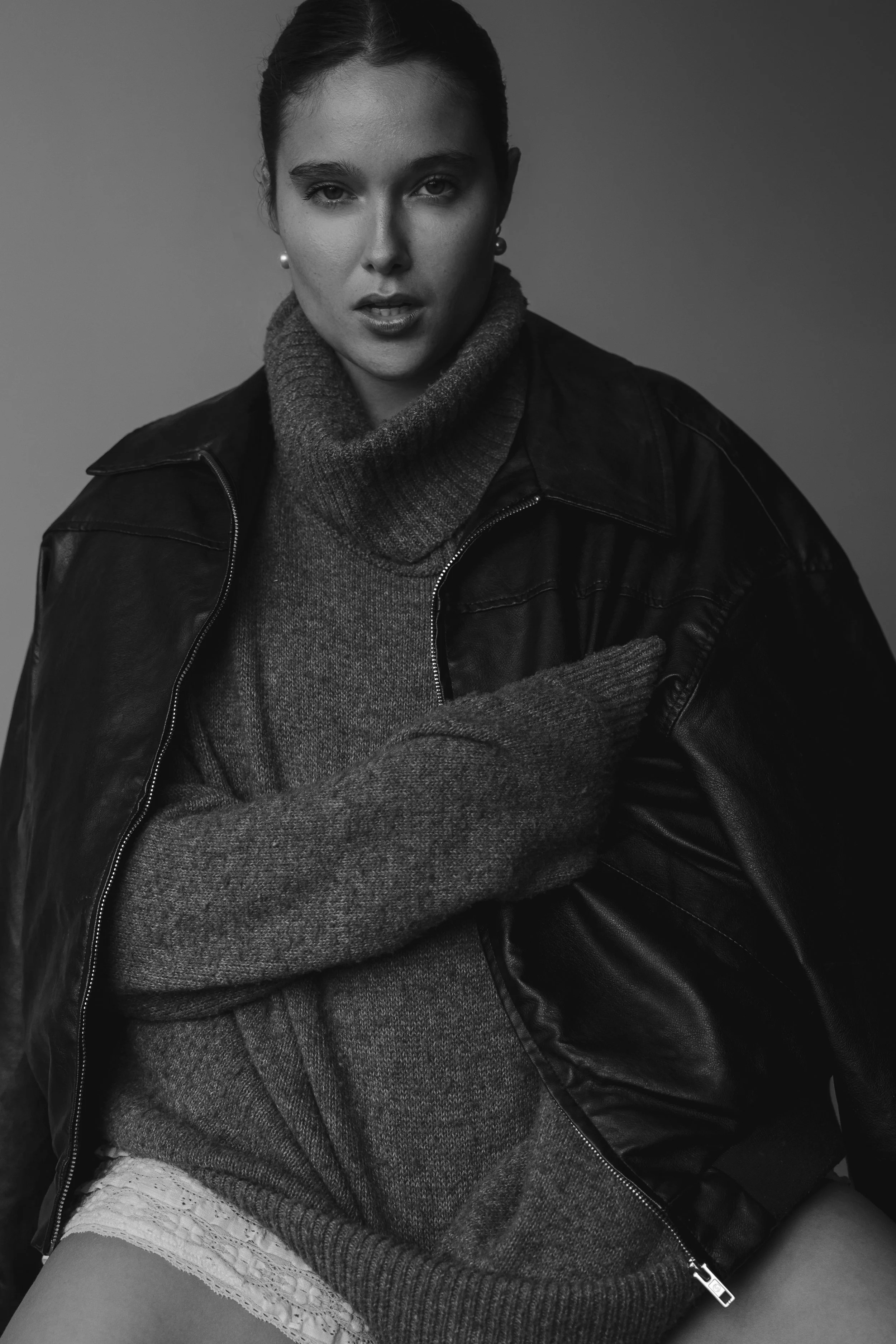 Black and white photograph of a woman with dark hair pulled back, wearing pearl earrings, a turtleneck sweater, and a leather jacket, with crossed arms and a serious expression.