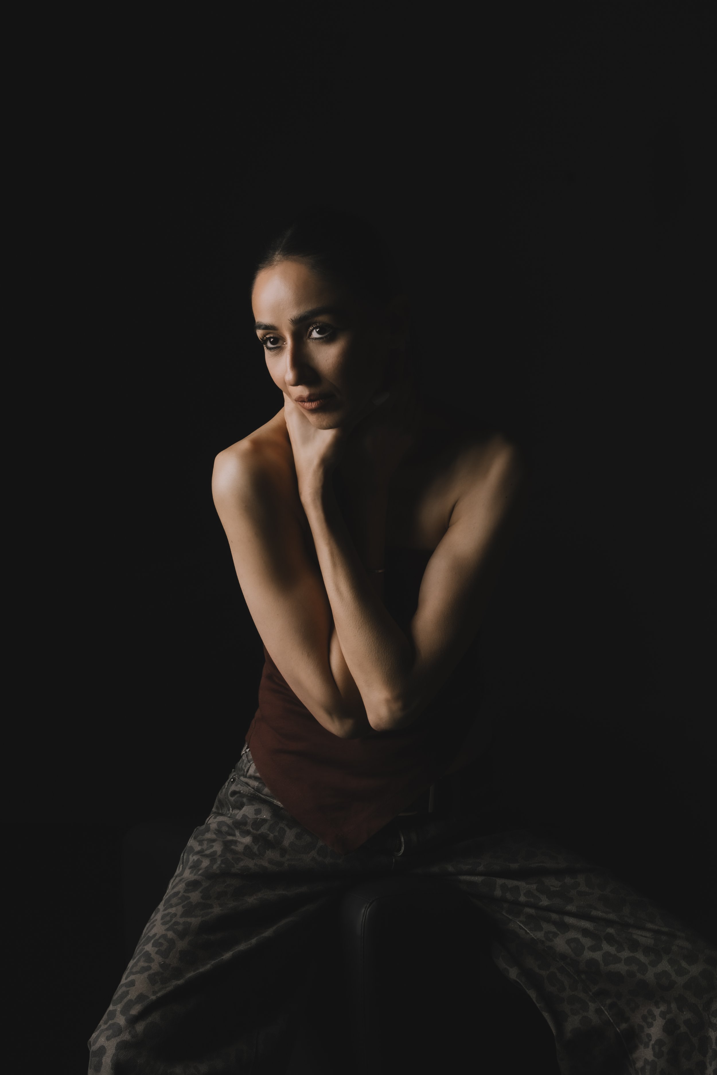 A woman with dark hair, makeup, and tan skin poses against a black background, wearing a dark top and camouflage pants, resting her chin on her hand.