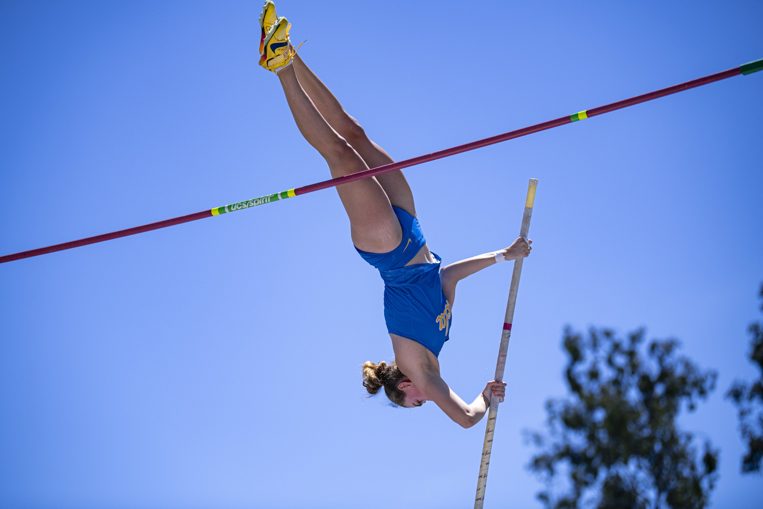 A female pole vaulter in a blue sports uniform is in mid-air, holding a pole and reaching for the bar during a competition against a clear blue sky.
