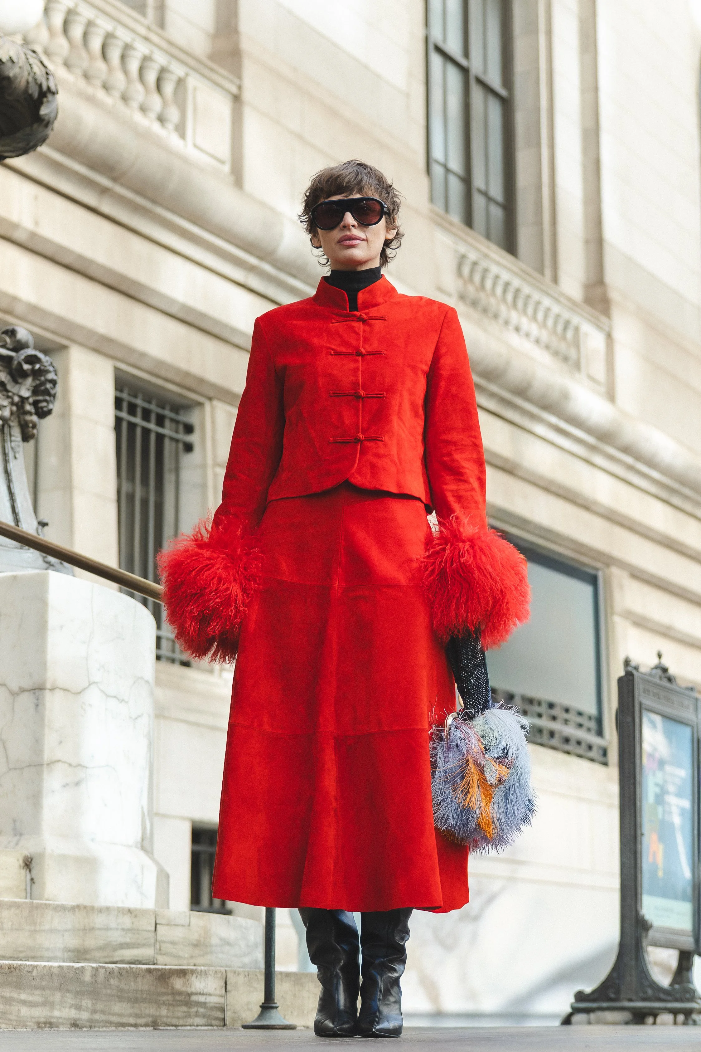 A woman wearing a red coat with oversized fluffy red cuffs, large sunglasses, and black boots, standing outdoors in front of a historic building with large windows and ornate architectural details.