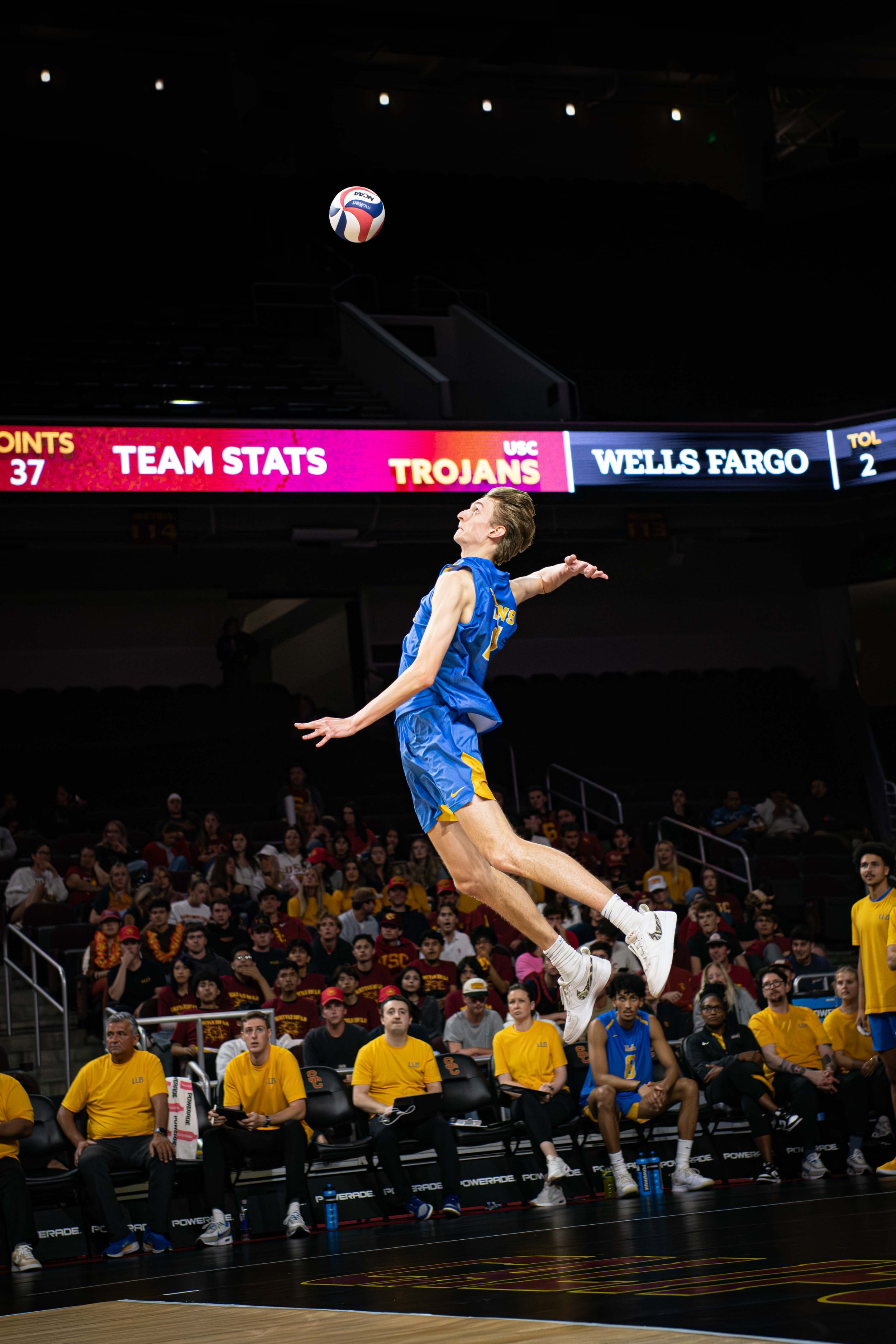 A female volleyball player in a blue uniform jumps to hit the ball during a match with spectators and team members seated courtside.