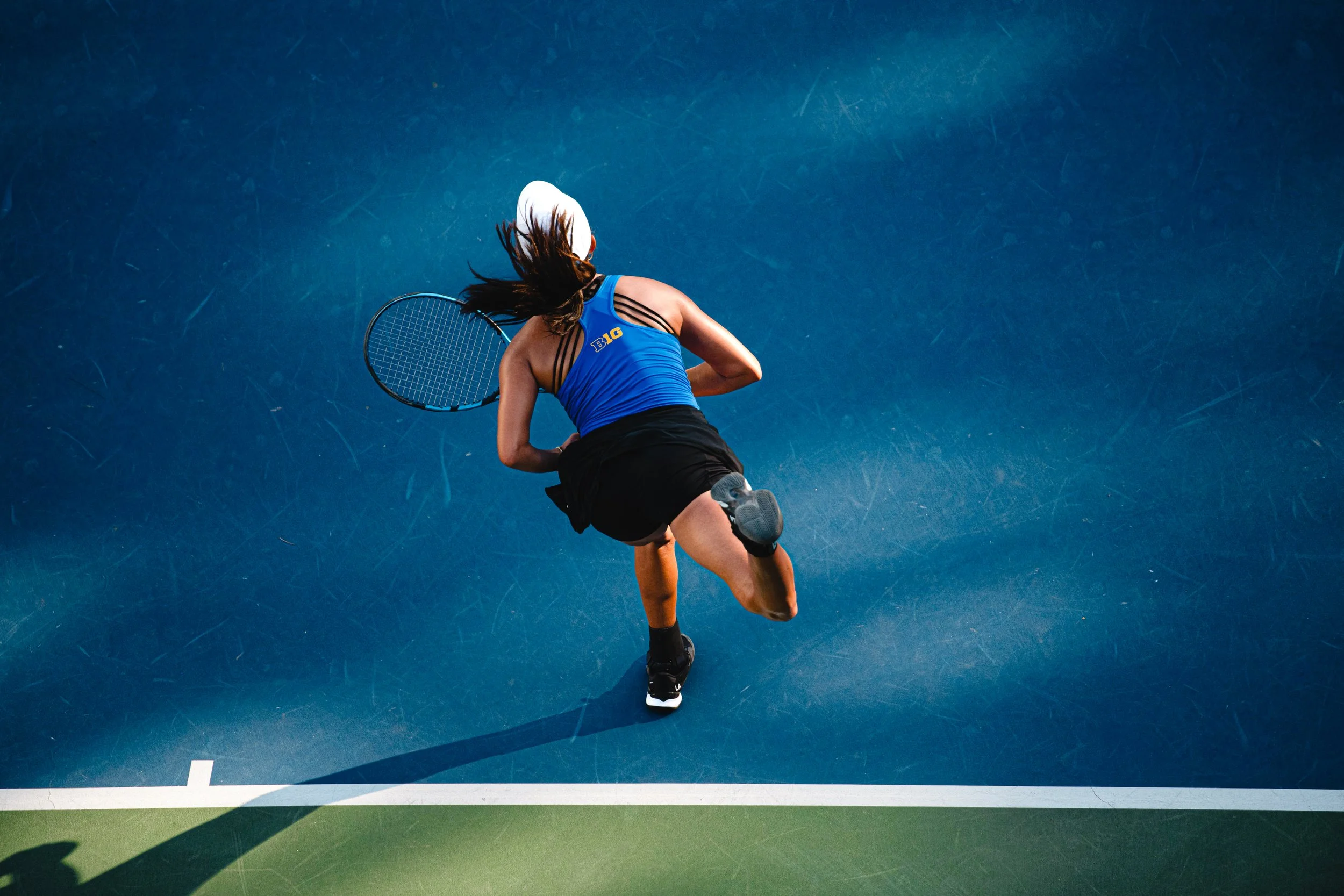 An overhead view of a female tennis player in a blue top and black shorts, swinging a tennis racket on a blue and green court.