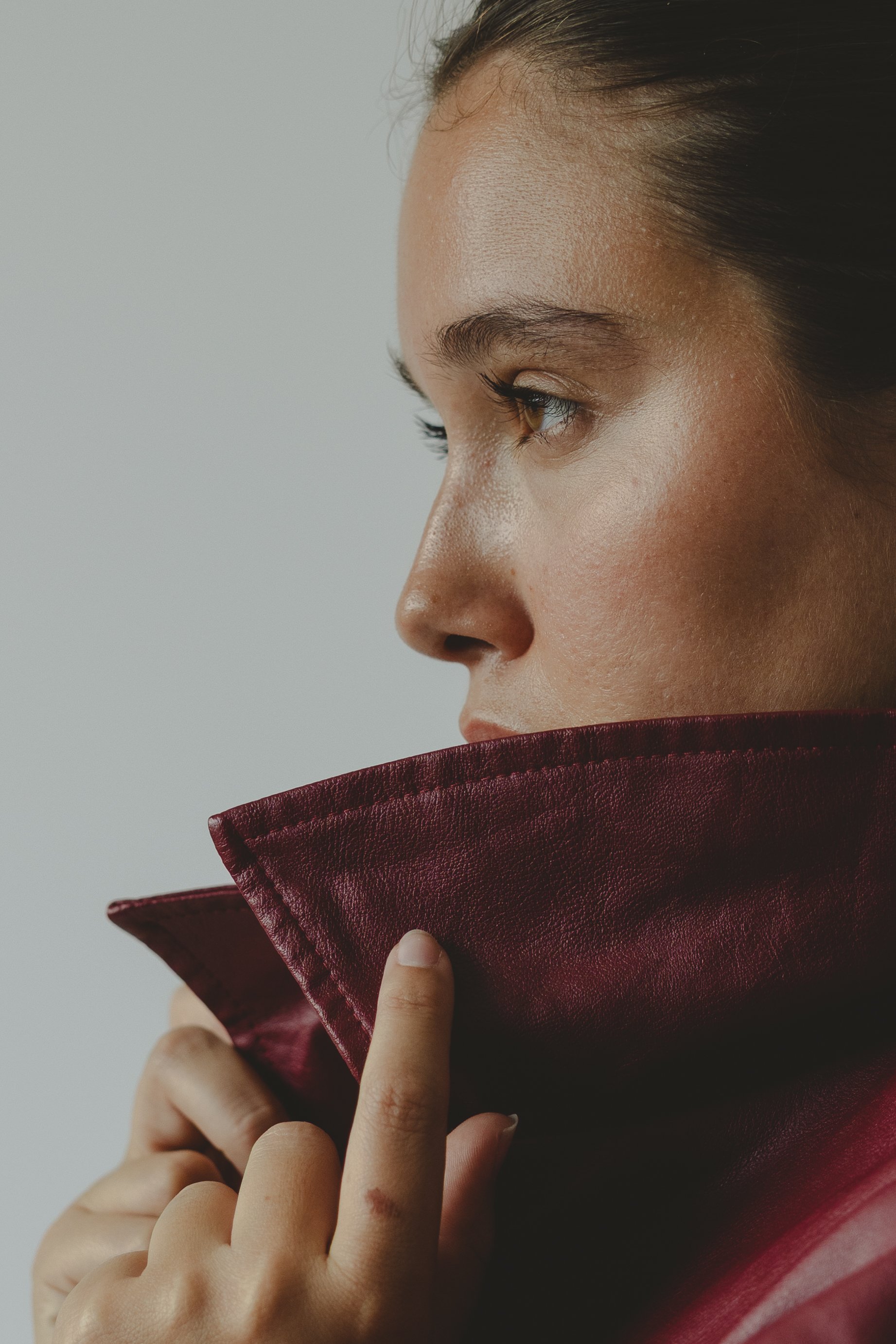 Close-up of a woman's face in profile, with her hand holding a burgundy leather garment near her chin, against a plain light background.