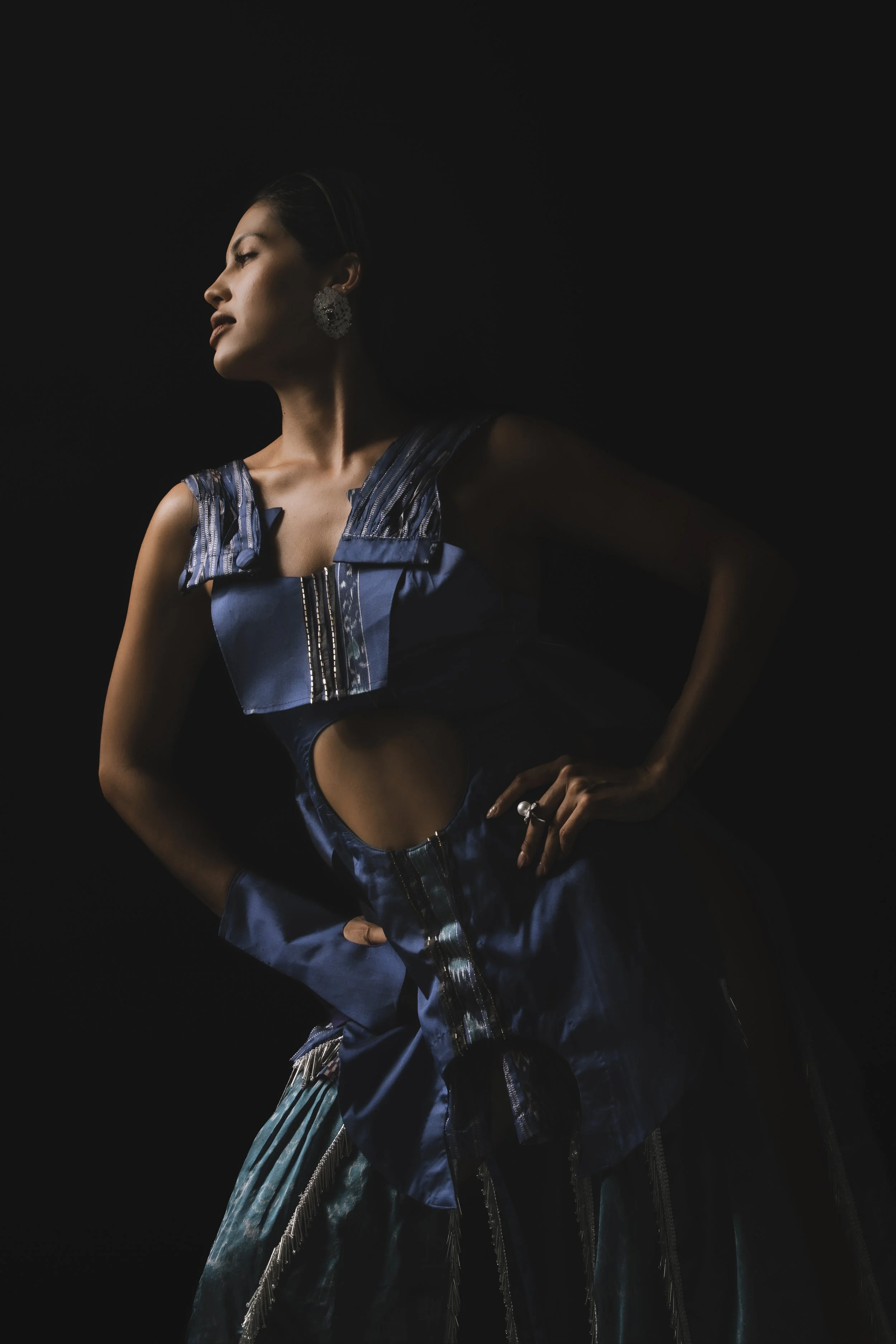 A woman wearing a metallic blue dress with fringe details and large earrings, posing with her eyes closed against a dark background.