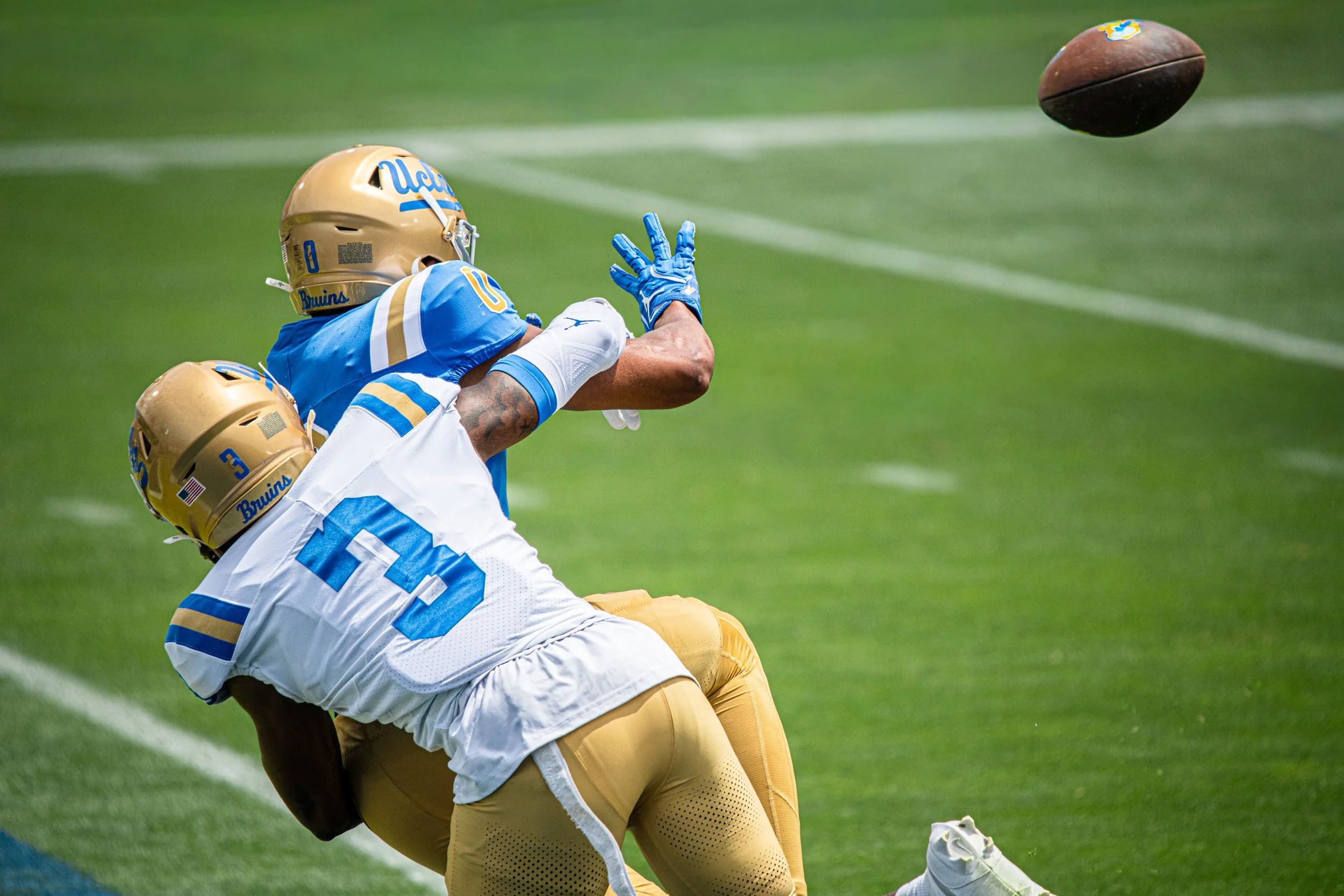 Two football players, one in a UCLA Bruins uniform, are on the ground trying to catch or block a football in midair during a game.