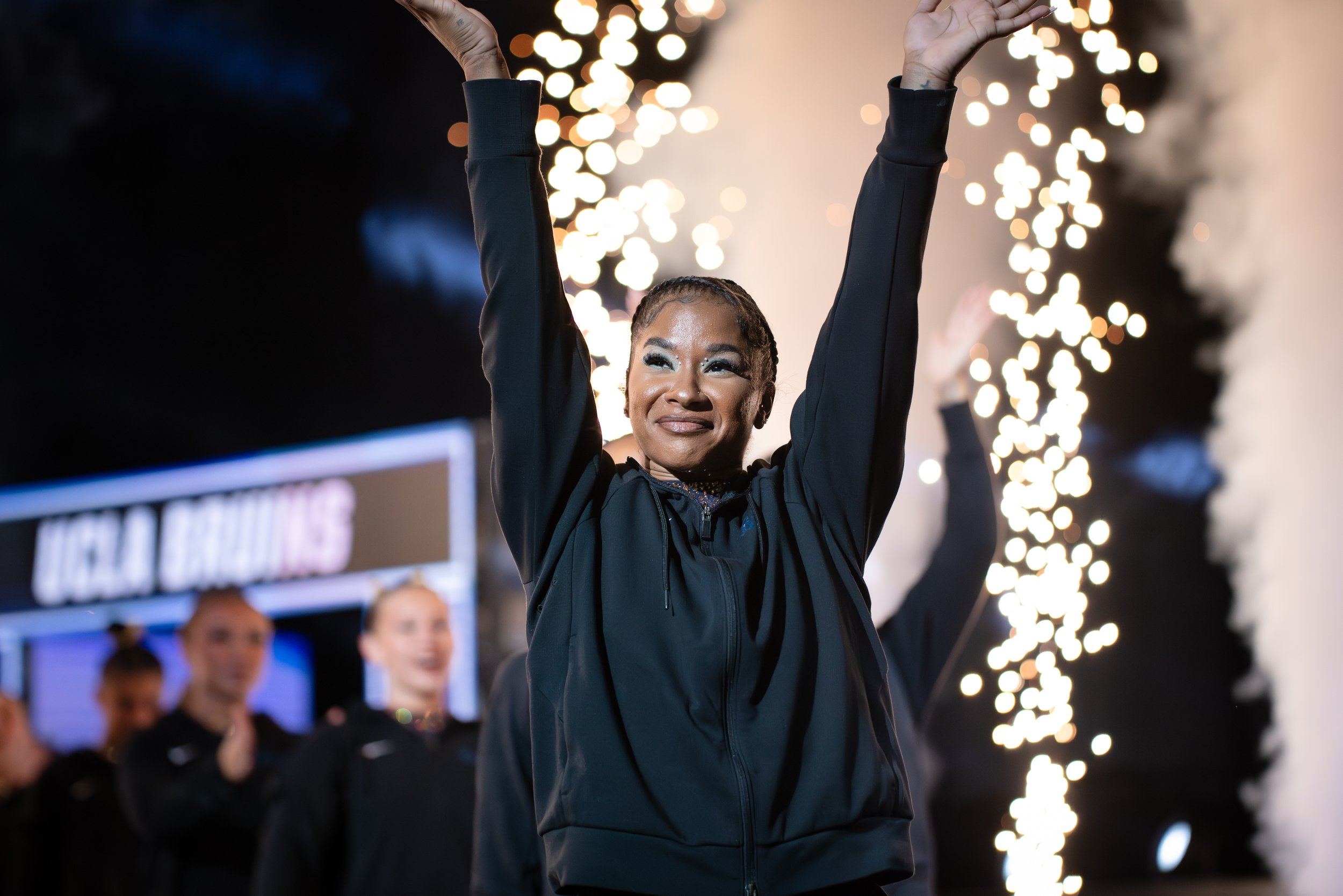 A woman with braided hair celebrating, raising her hands in the air, surrounded by sparklers and blurred faces at a nighttime event.