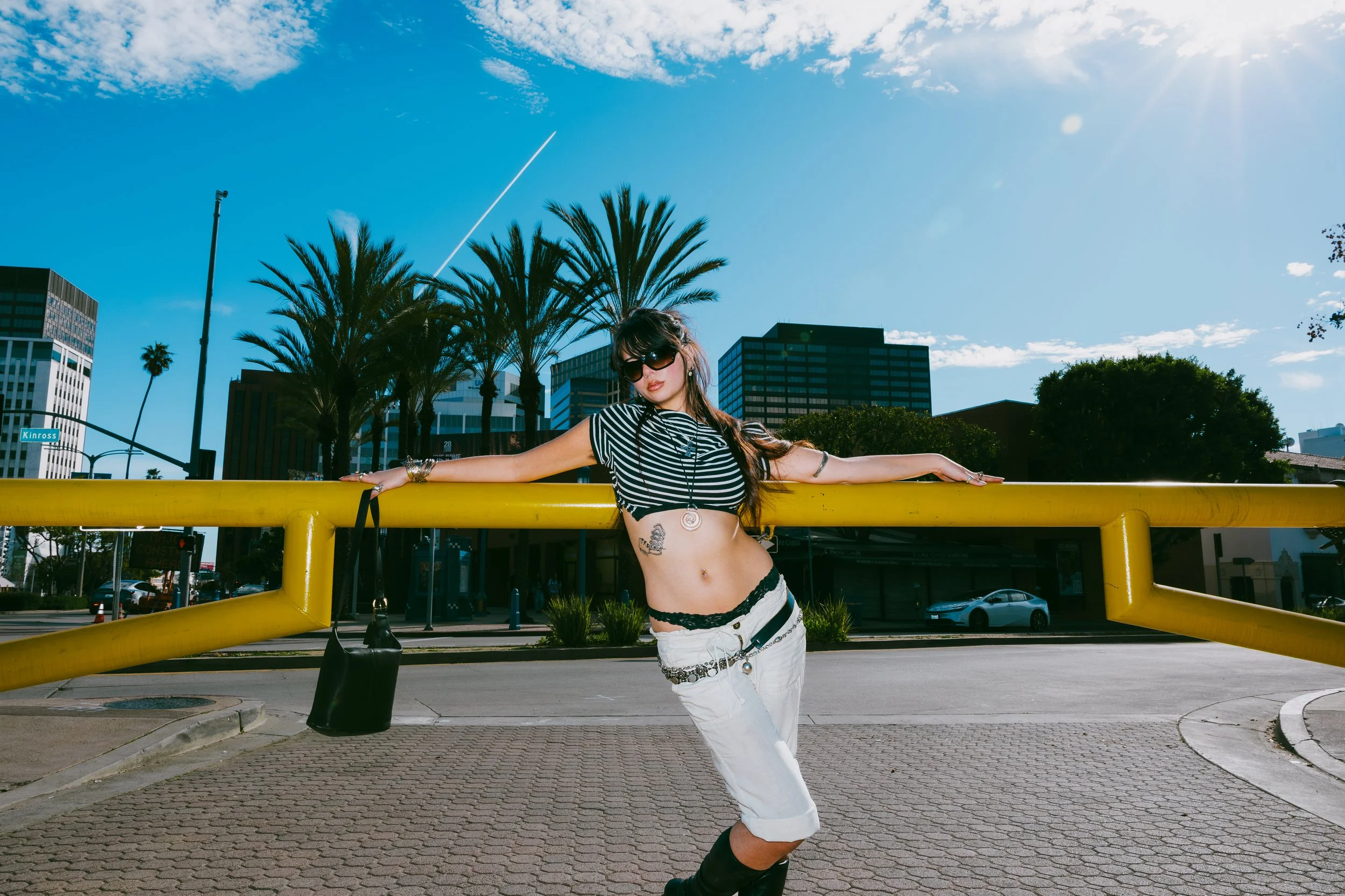 Woman with long dark hair wearing sunglasses, a black and white striped crop top, and white pants with black lace details, posing with arms outstretched on a yellow barrier in an urban area with palm trees, tall buildings, and a clear blue sky.