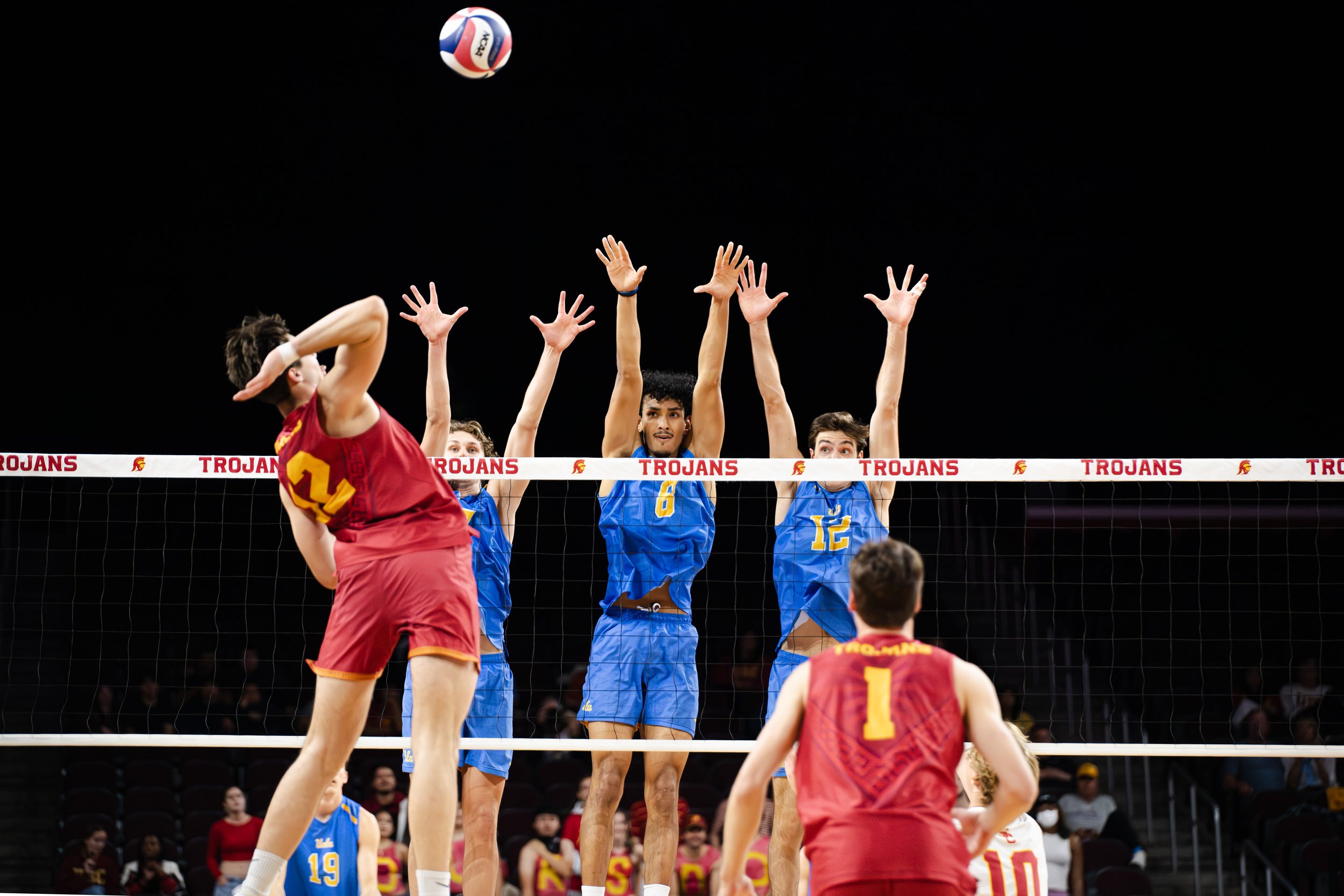 A volleyball game with players in red and blue uniforms. The player in red is spiking the ball, while players in blue attempt to block. The volleyball is in the air above the net, and the net has the word 'TROJANS' on it.