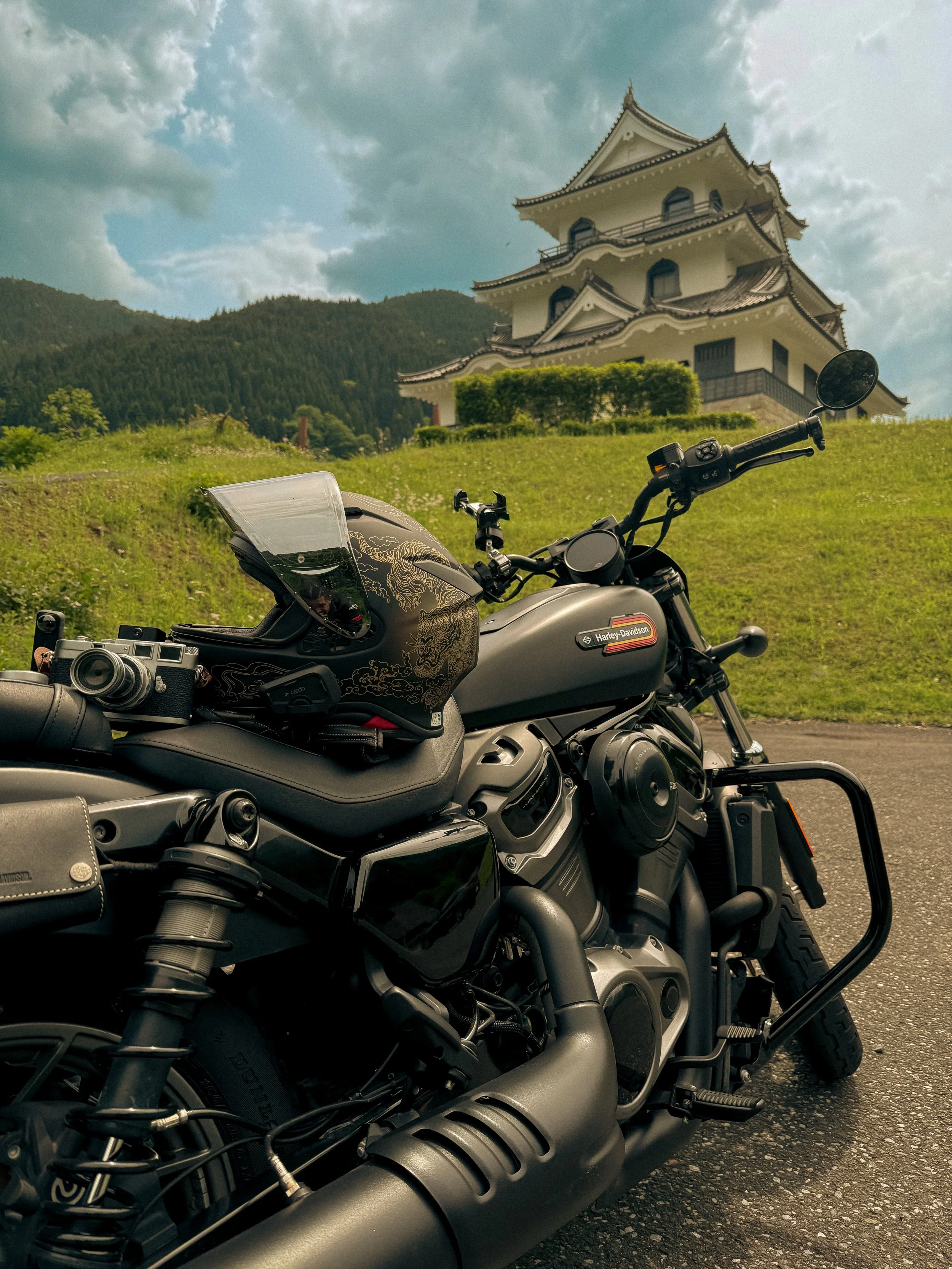 Close-up of a black Harley-Davidson motorcycle with a decorative helmet and gear on the seat, parked on a paved surface in front of a traditional Japanese castle and green hillside under a partly cloudy sky.