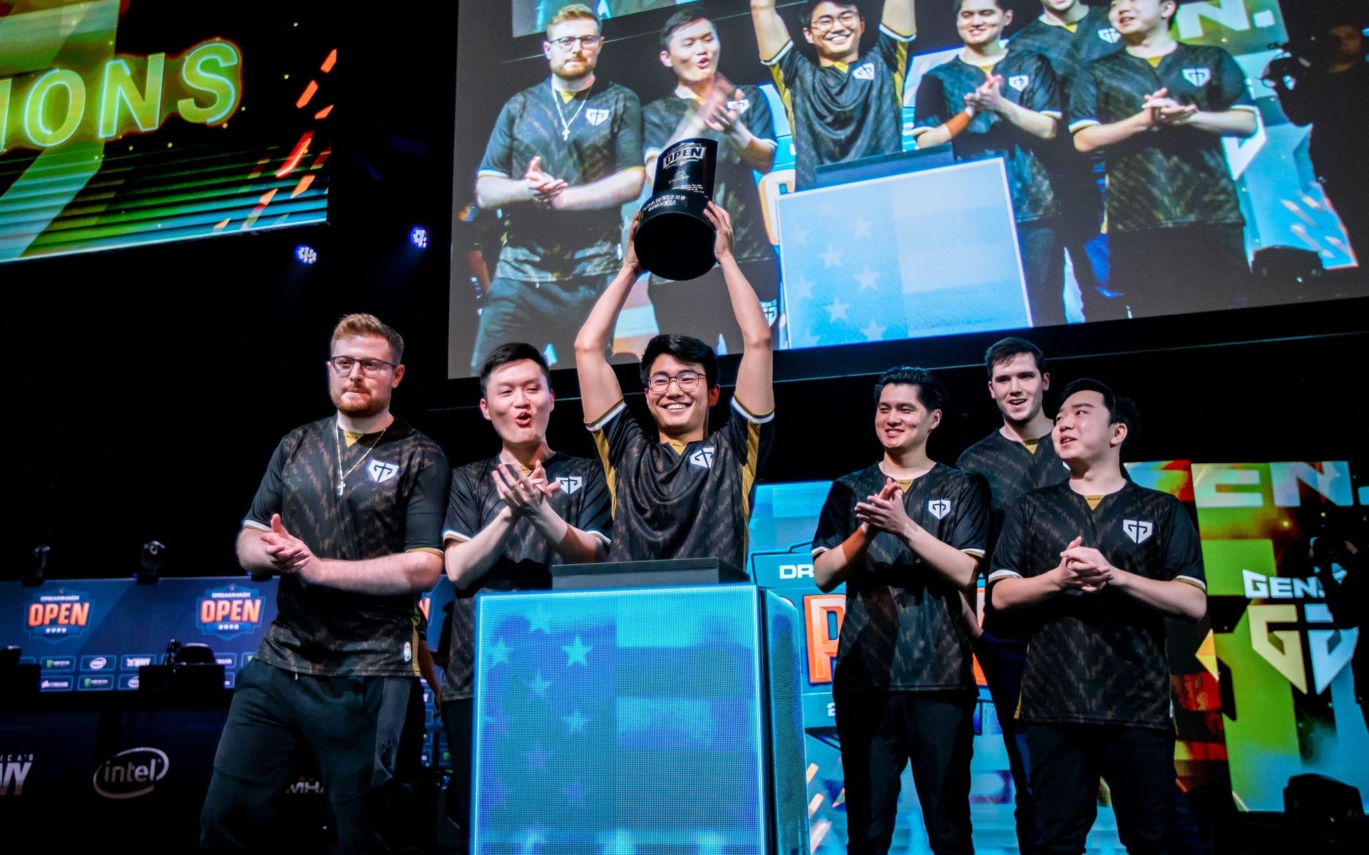 A team of six esports players celebrating on stage with one holding a trophy above his head, during an event with large screens and colorful banners in the background.
