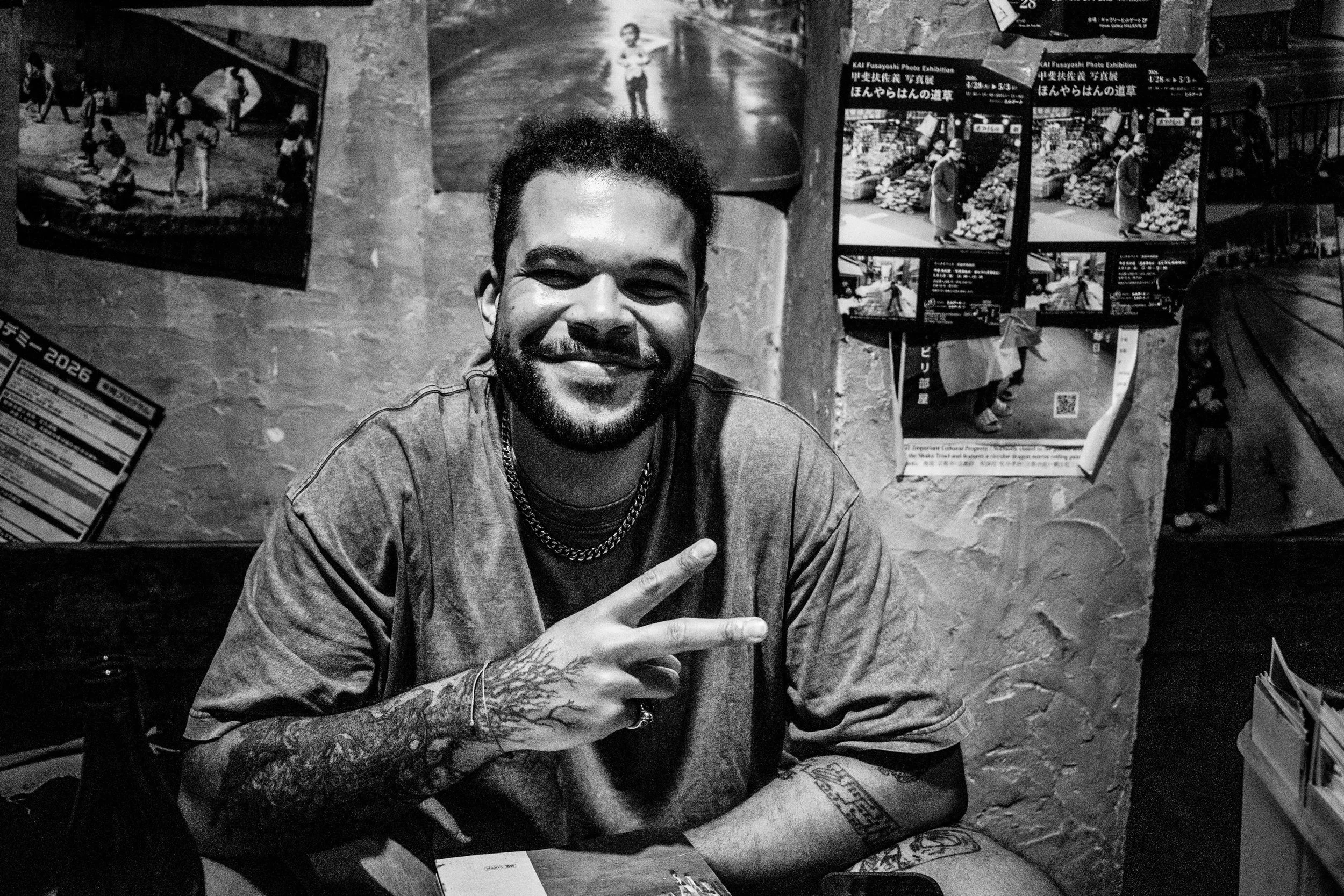A smiling man with a beard and tattoos on his arm, making a peace sign with his hand. He is sitting at a table with posters and photographs on the wall behind him.
