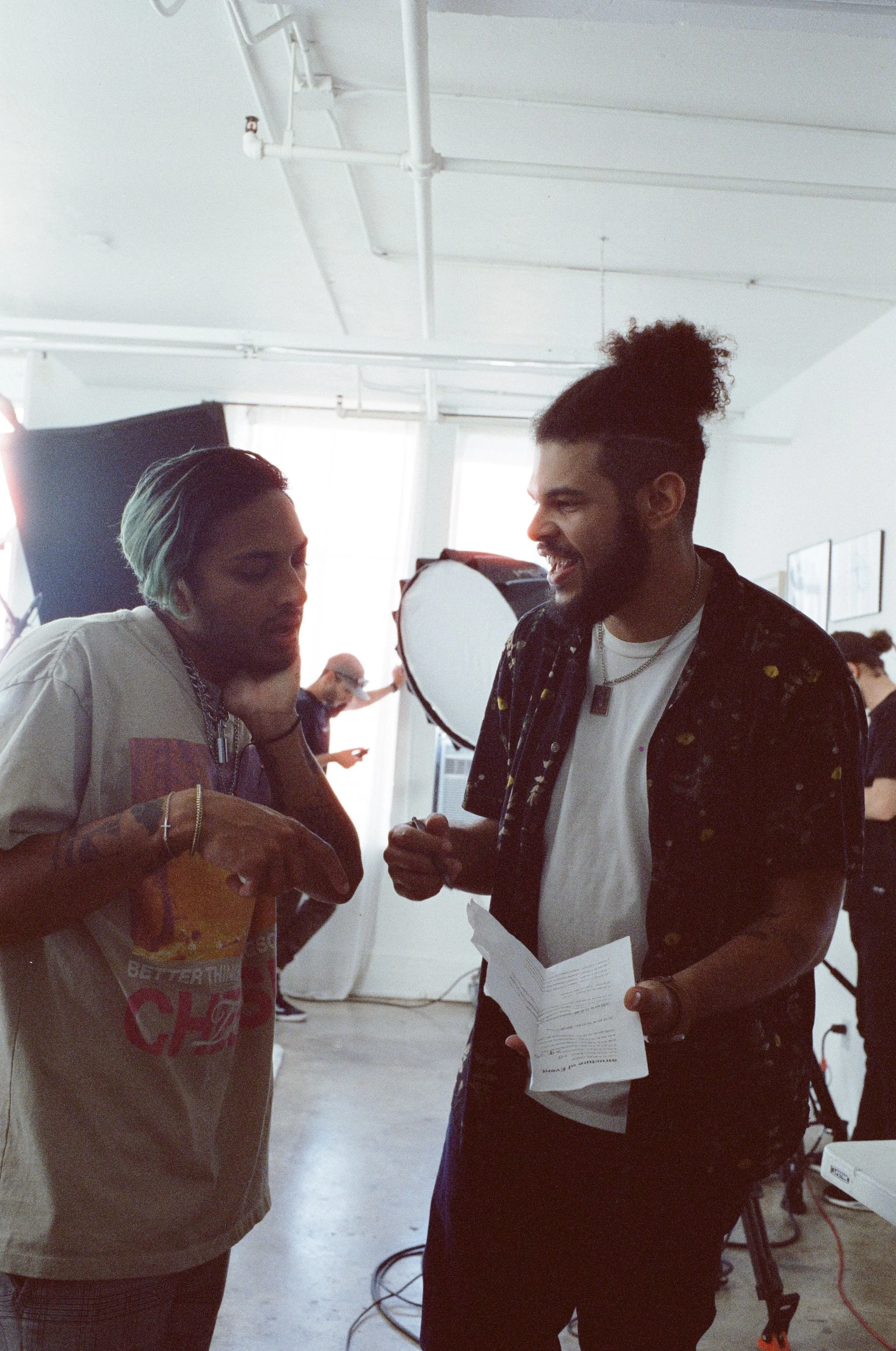 Two people having a conversation in a studio setting. The person on the left has short blonde hair and tattoos, holding a bag and looking at the person on the right. The person on the right has curly hair tied up in a bun, a beard, and is holding papers, smiling, and wearing layered necklaces.