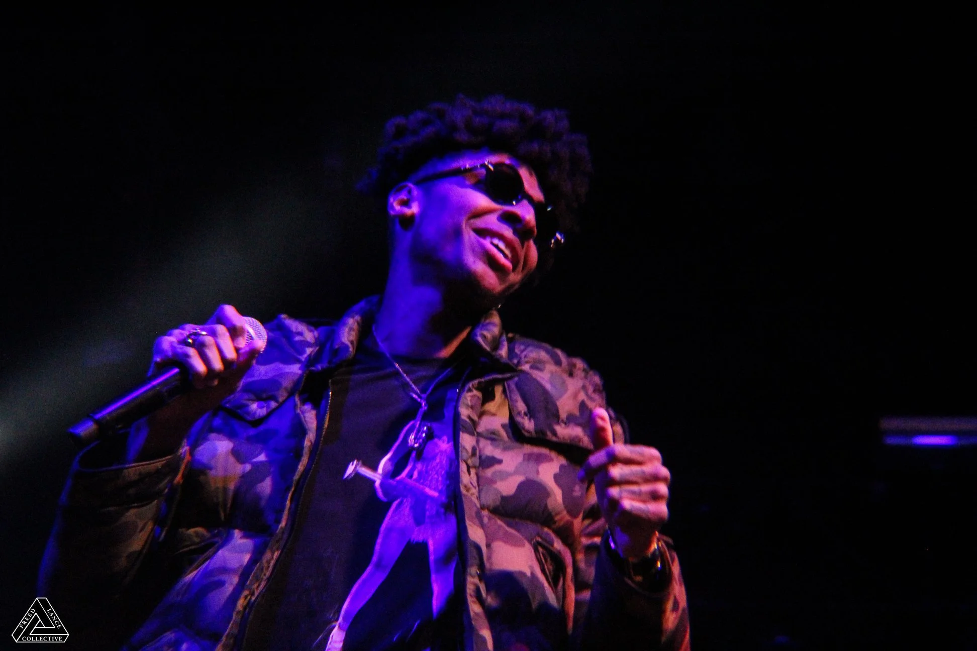 A person with dark skin, wearing sunglasses and a camouflage jacket, smiling and holding a microphone in a dark environment with purple lighting.