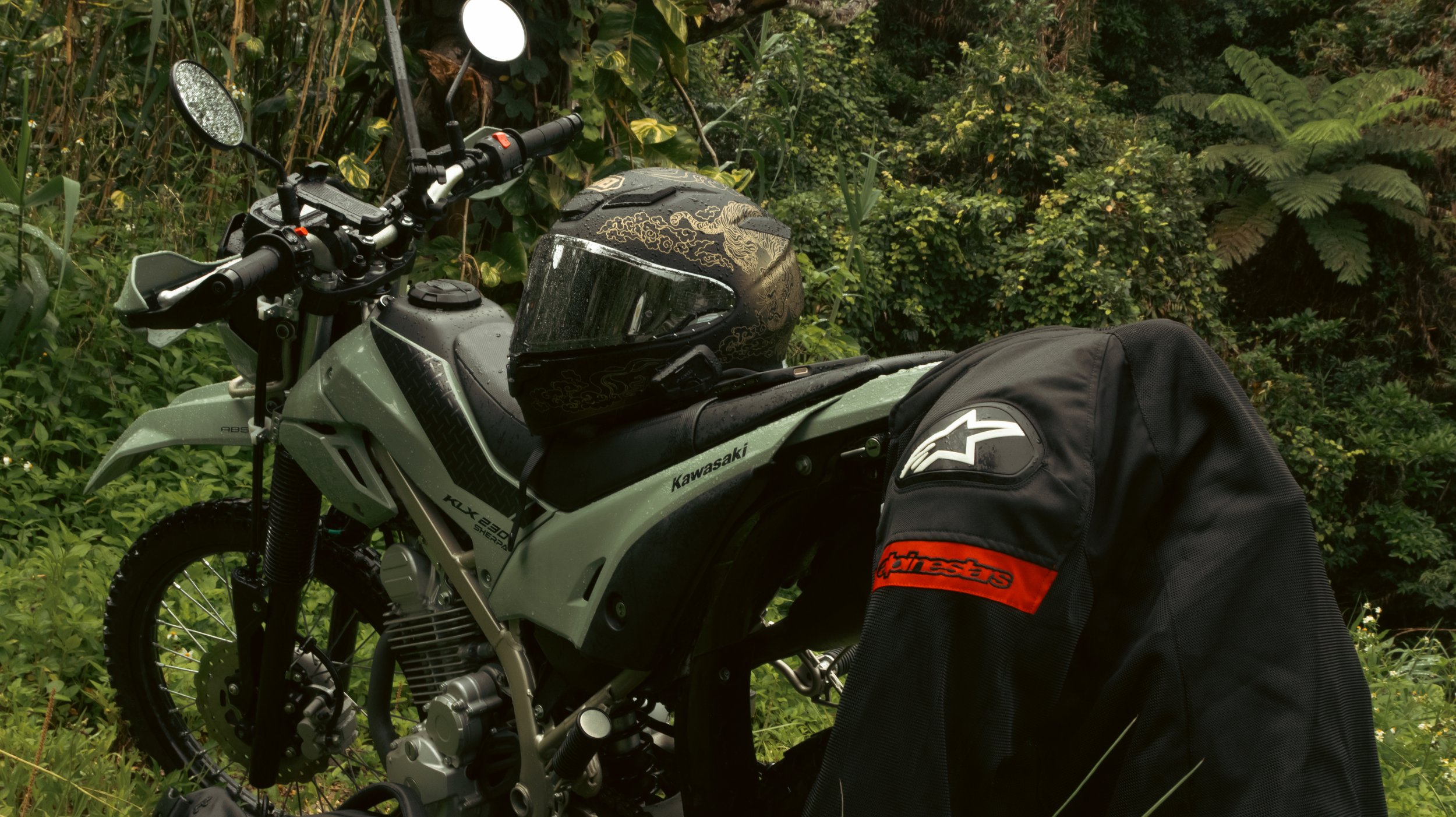 A Kawasaki off-road motorcycle equipped with a black and gold helmet on the seat, a black Alpinestars backpack attached to the bike, and rain droplets on the helmet visor, set against a lush green, foliage-filled background.