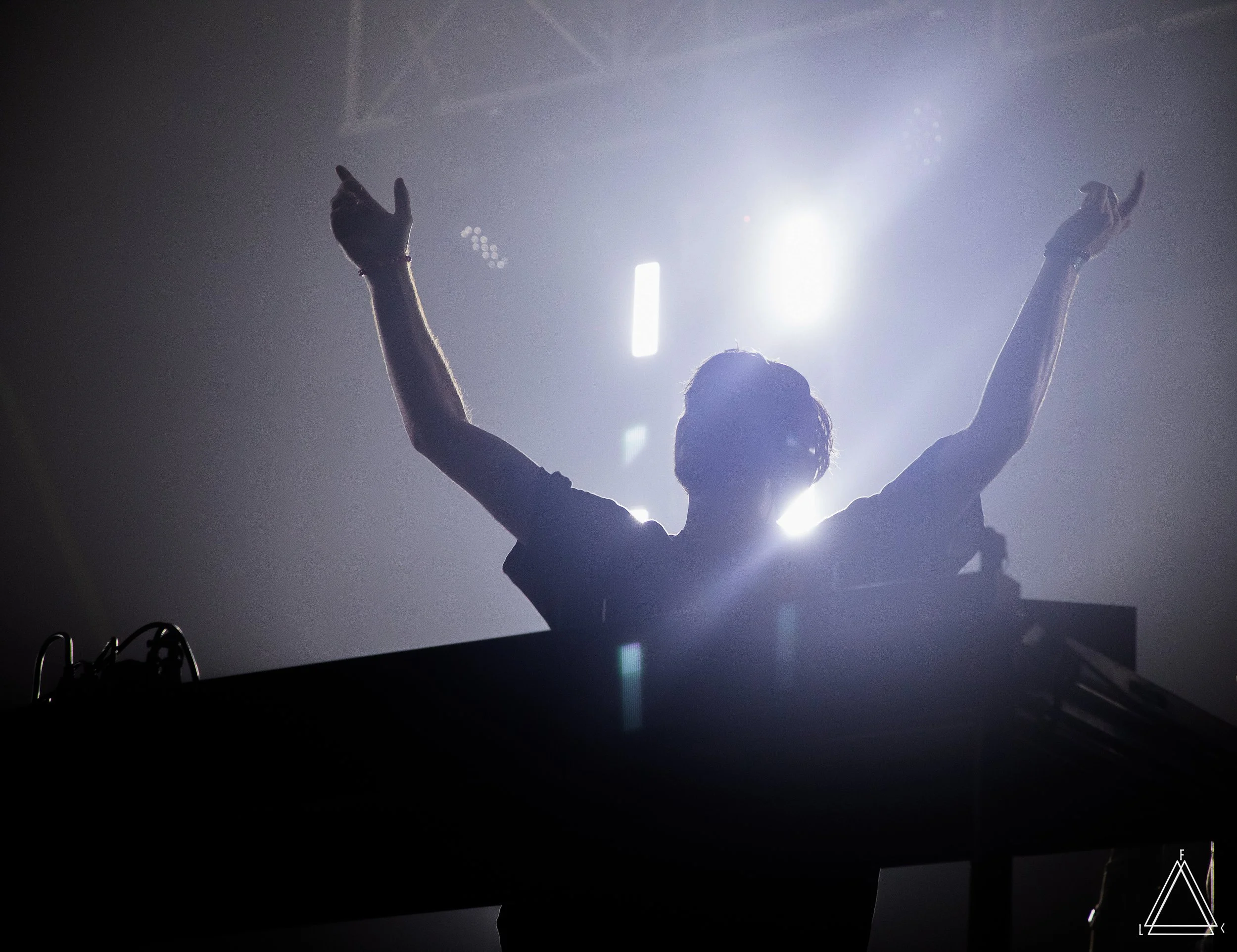 Person with arms raised performing on stage under bright lights, dark background, and stage equipment visible at the bottom.