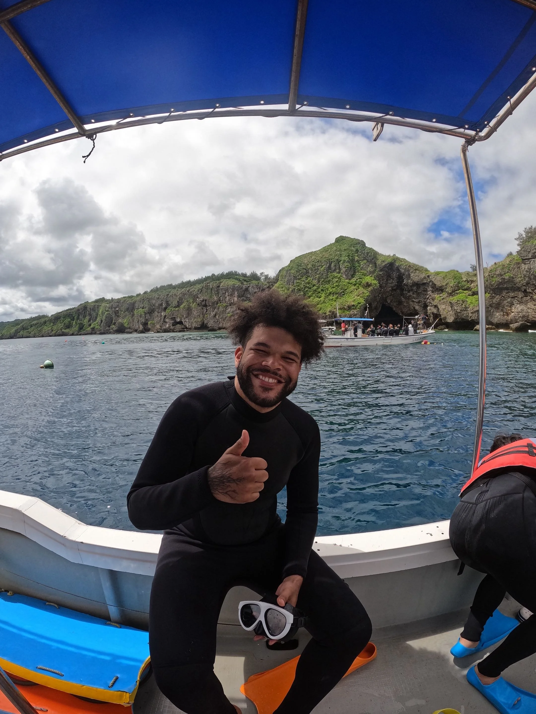 A man smiling and giving a thumbs-up on a boat, holding snorkeling mask and surrounded by water, with a rocky green island and another boat in the background.