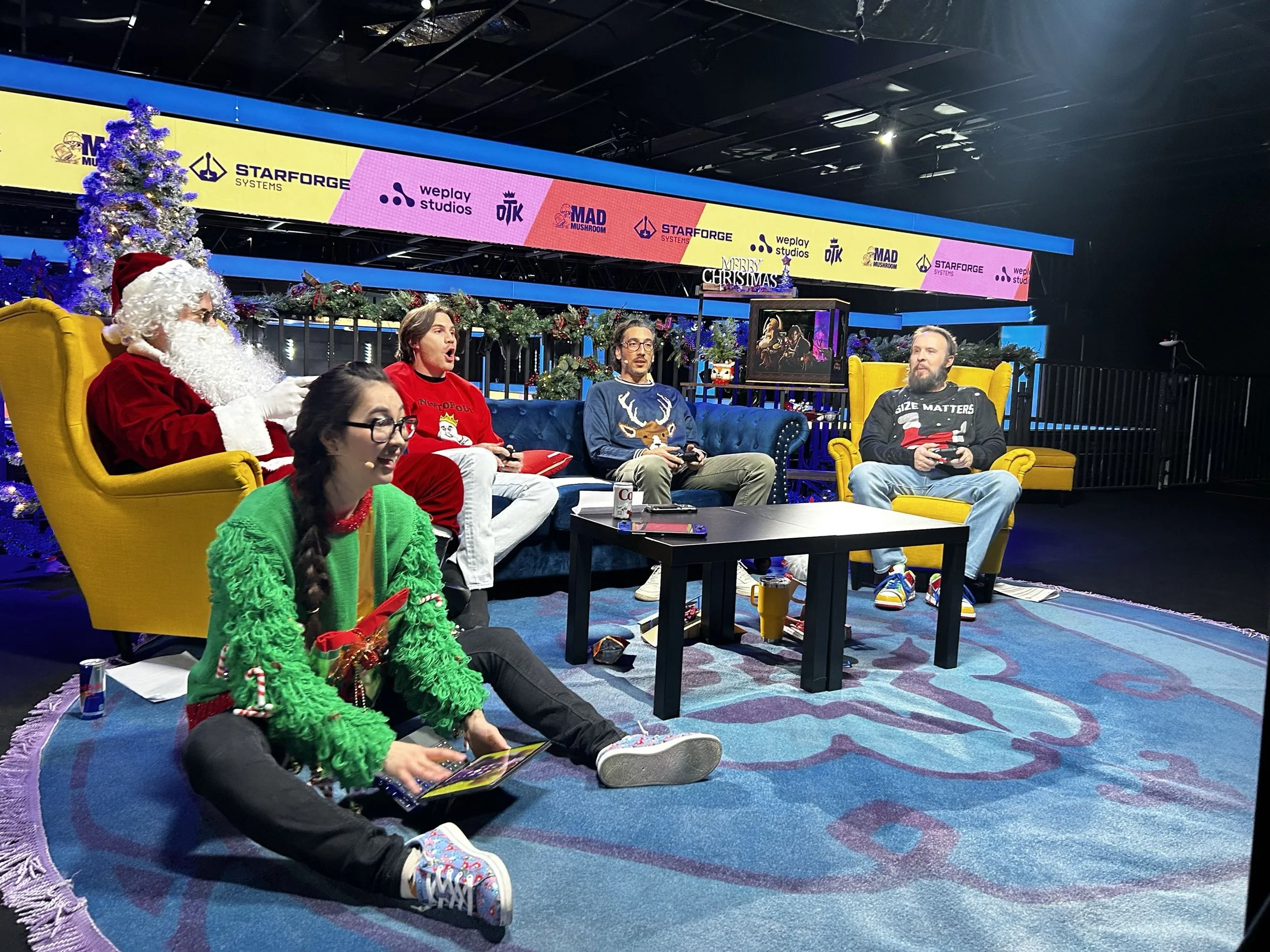 A holiday gathering with several people sitting and on the floor, some dressed in Christmas sweaters and one person dressed as Santa Claus, in a festive decorated room with Christmas trees and garlands. There is a large screen displaying sponsor logos behind them.