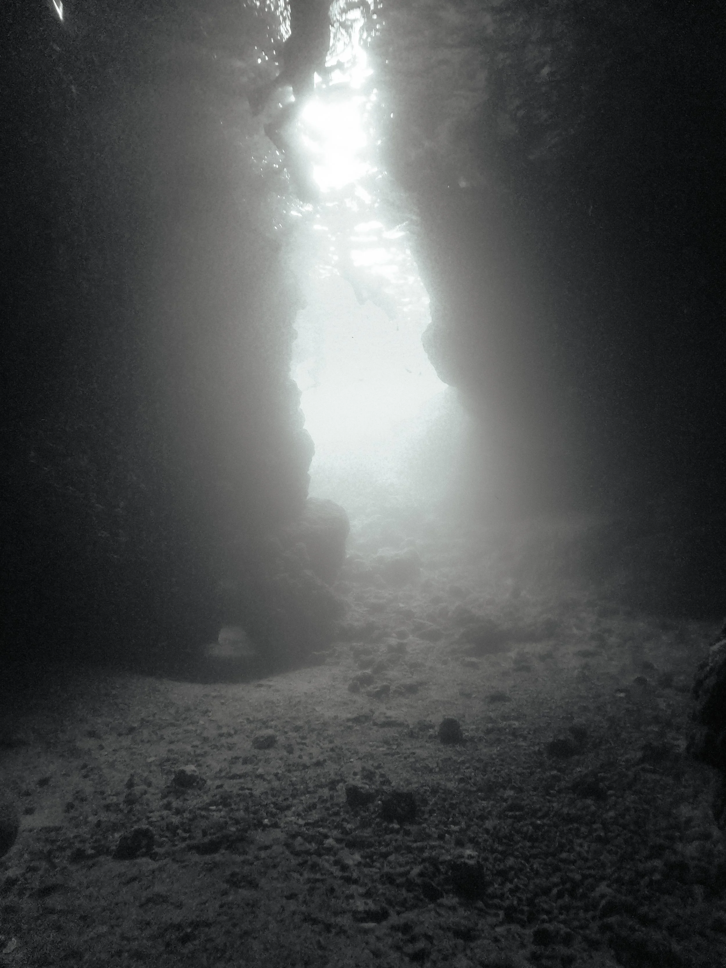 A dark underwater scene featuring a narrow crevice with light shining through from above, illuminating the seabed.