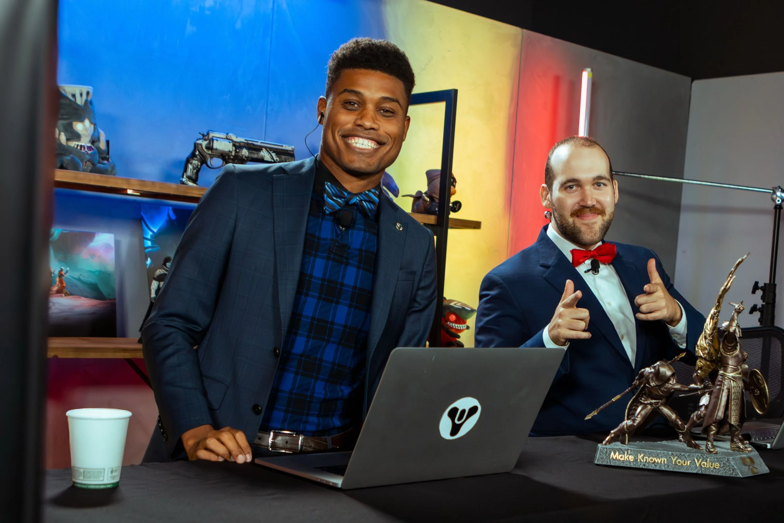 Two men dressed in suits, one wearing a bow tie, sitting at a desk with a laptop and a Sci-Fi themed figurine. The background has colorful wall art and shelves with various objects. The men are smiling and posing for the camera.