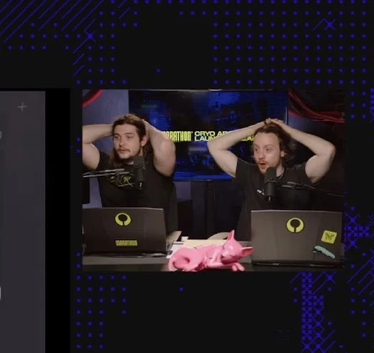 Two men sitting at a table in a studio with microphones and laptops, holding their heads with hands, appearing surprised or shocked. Pink plush toy on the table, background electronic equipment and screen.