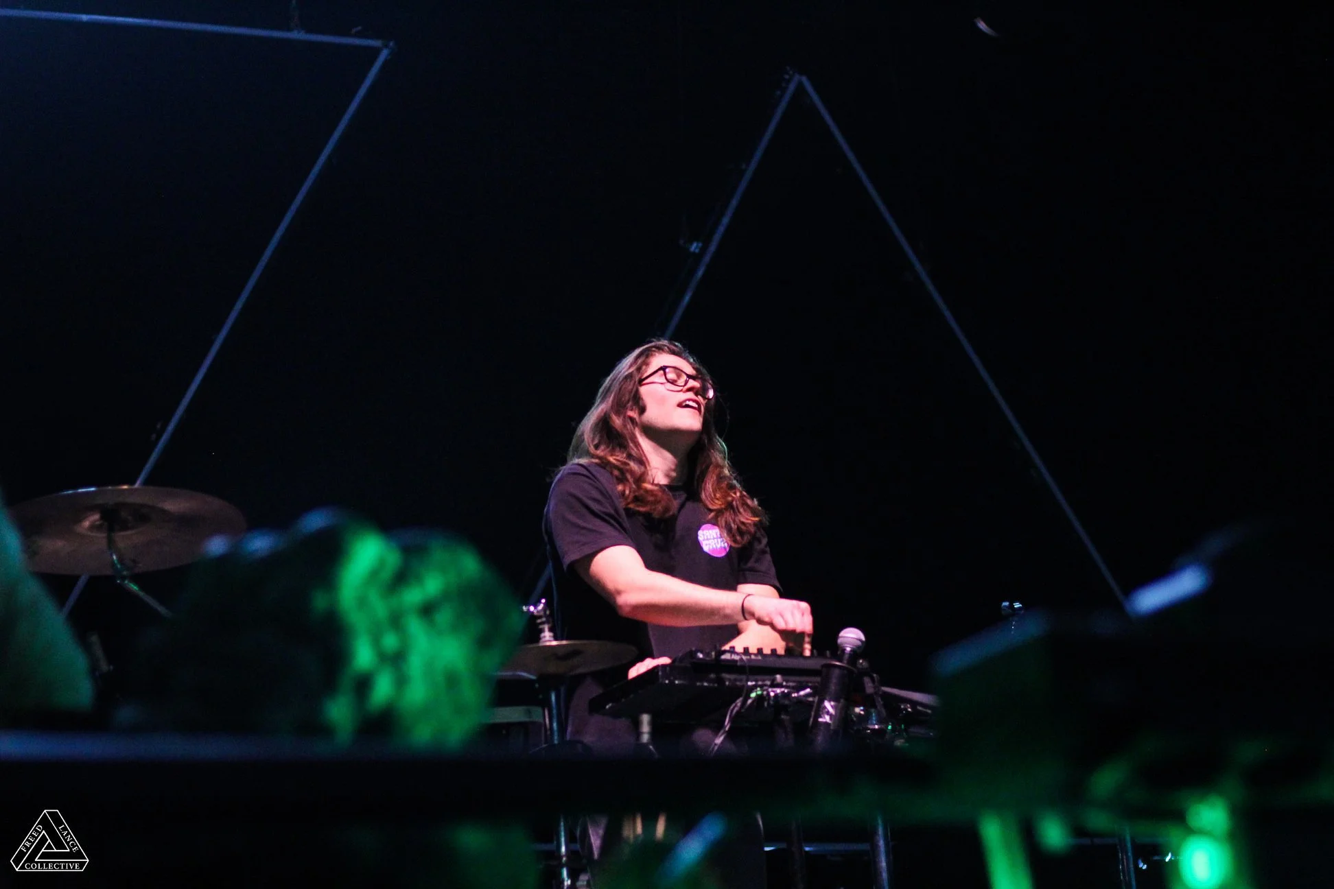 A woman with long hair and glasses DJing on stage with a dark background and some stage lighting, surrounded by a crowd.