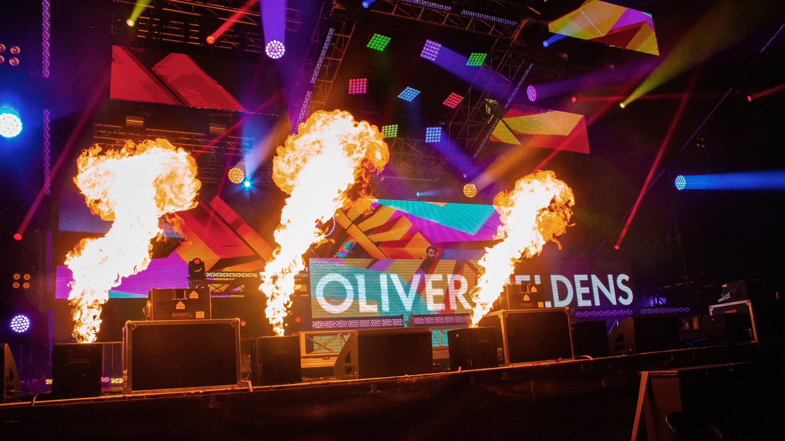 Stage with colorful lights, large screens, and pyrotechnics including flames shooting upward. The screens display the name Oliver Dens.