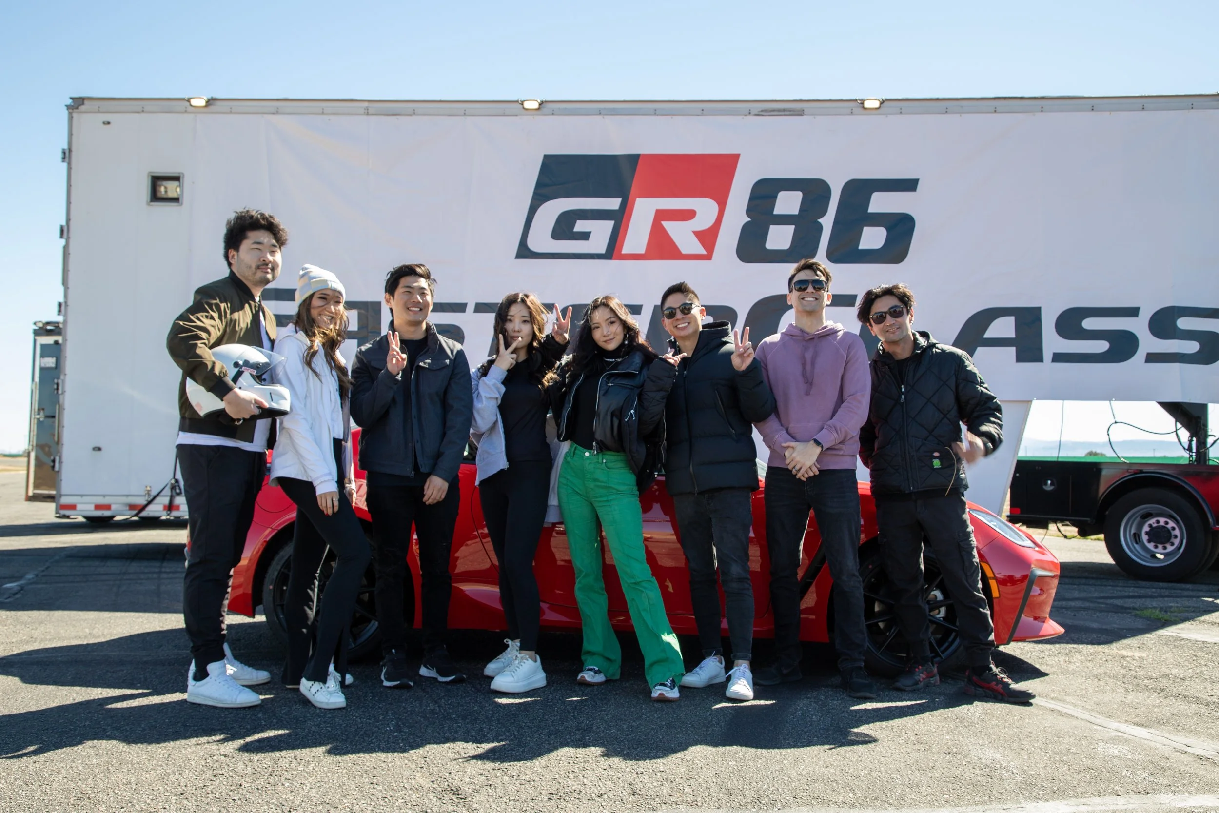 Group of people standing in front of a racing team truck with a red sports car. The group is smiling and making peace signs, dressed casually with some wearing sunglasses and racing gear. The truck has a racing logo that reads 'GR86.'