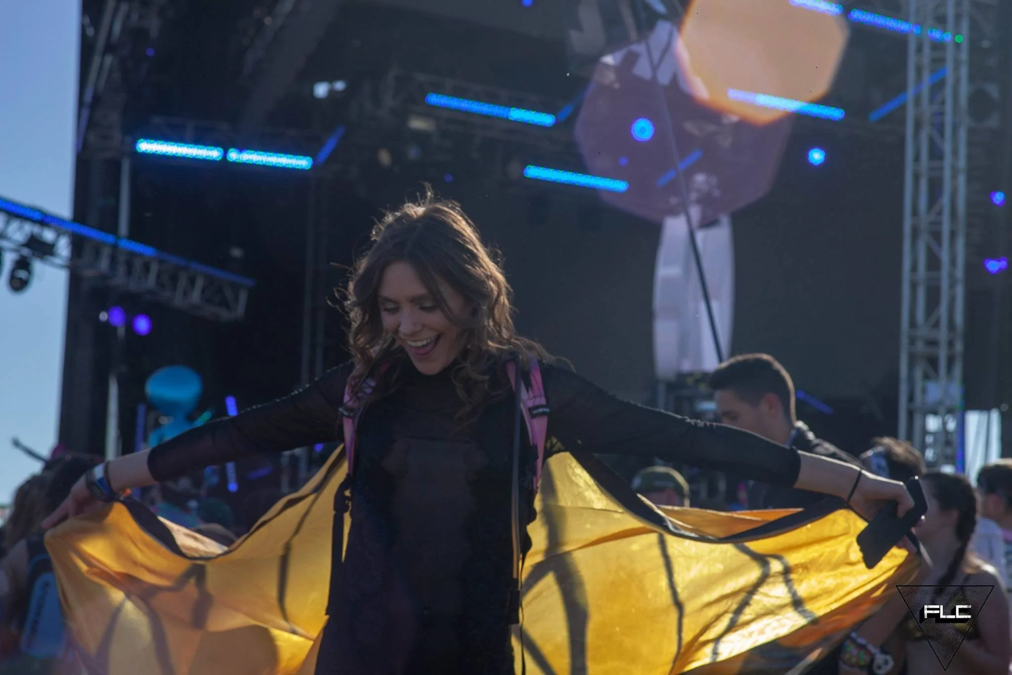 A woman standing outdoors on stage, smiling and holding a yellow parachute or canopy, with a large stage and lighting in the background, during what appears to be a public event or celebration.