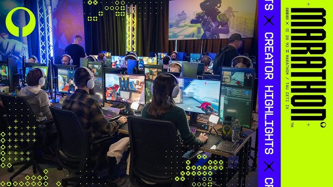 Multiple people sitting at desks with gaming computers and wearing headphones in a gaming event or tournament setting, with large screens displaying games and a promotional banner for a gaming event.