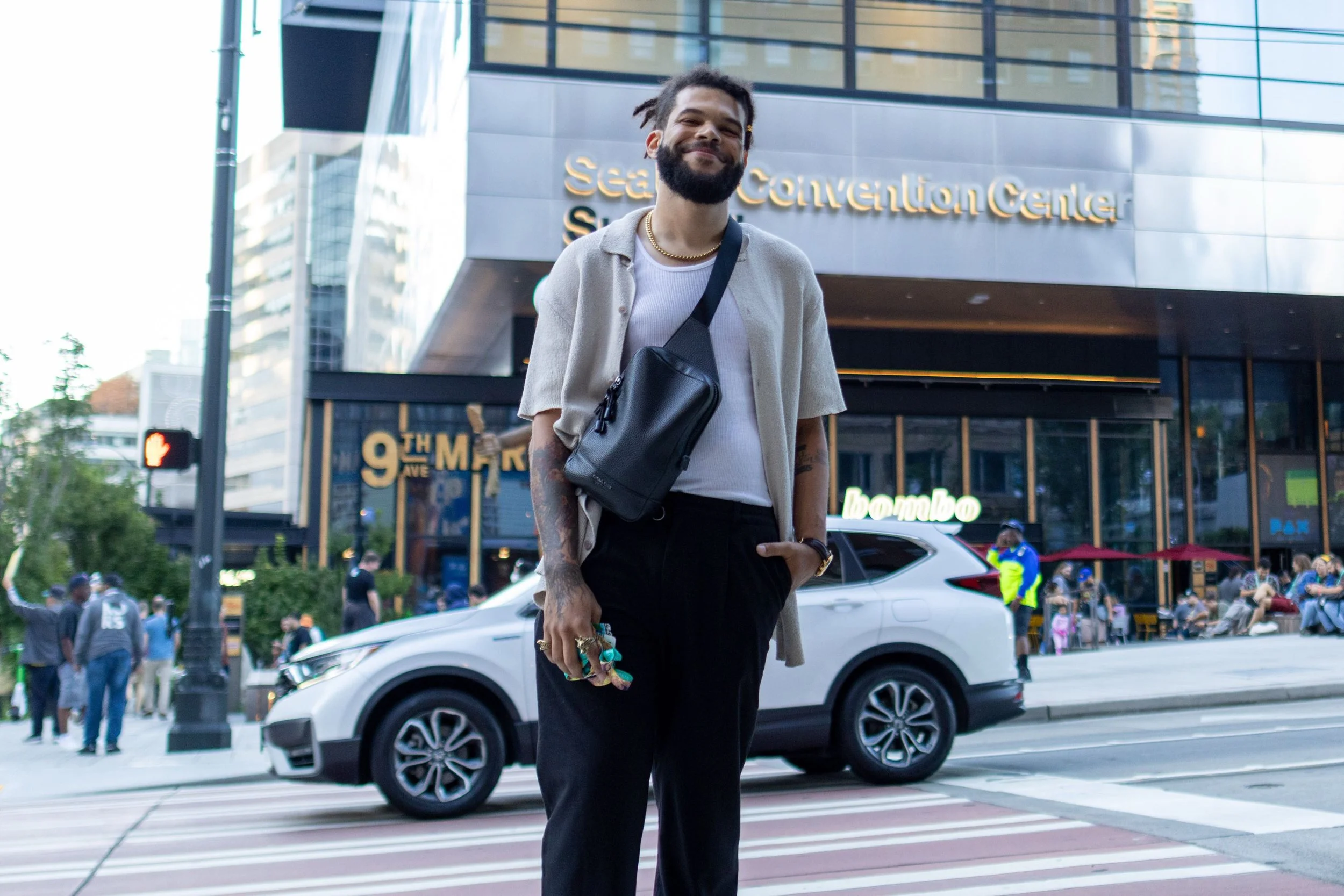 A man with a beard and dreadlocks smiling in front of the Seattle Convention Center. He is wearing a beige jacket, white shirt, black pants, and has a small black bag across his shoulder. There are cars, people, and buildings in the background.