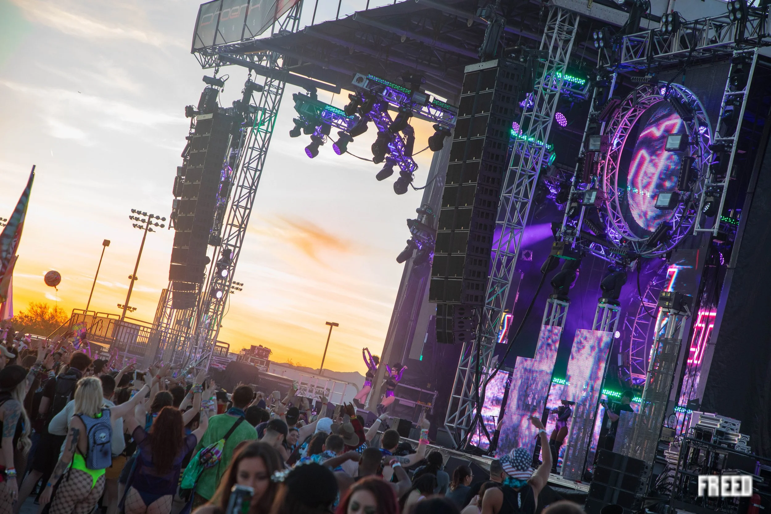 A large outdoor concert stage with vibrant purple and green lighting, filled with speakers, truss, and musical equipment. A crowd of people facing the stage, some raising hands and wearing colorful outfits, is visible in the foreground during sunset.