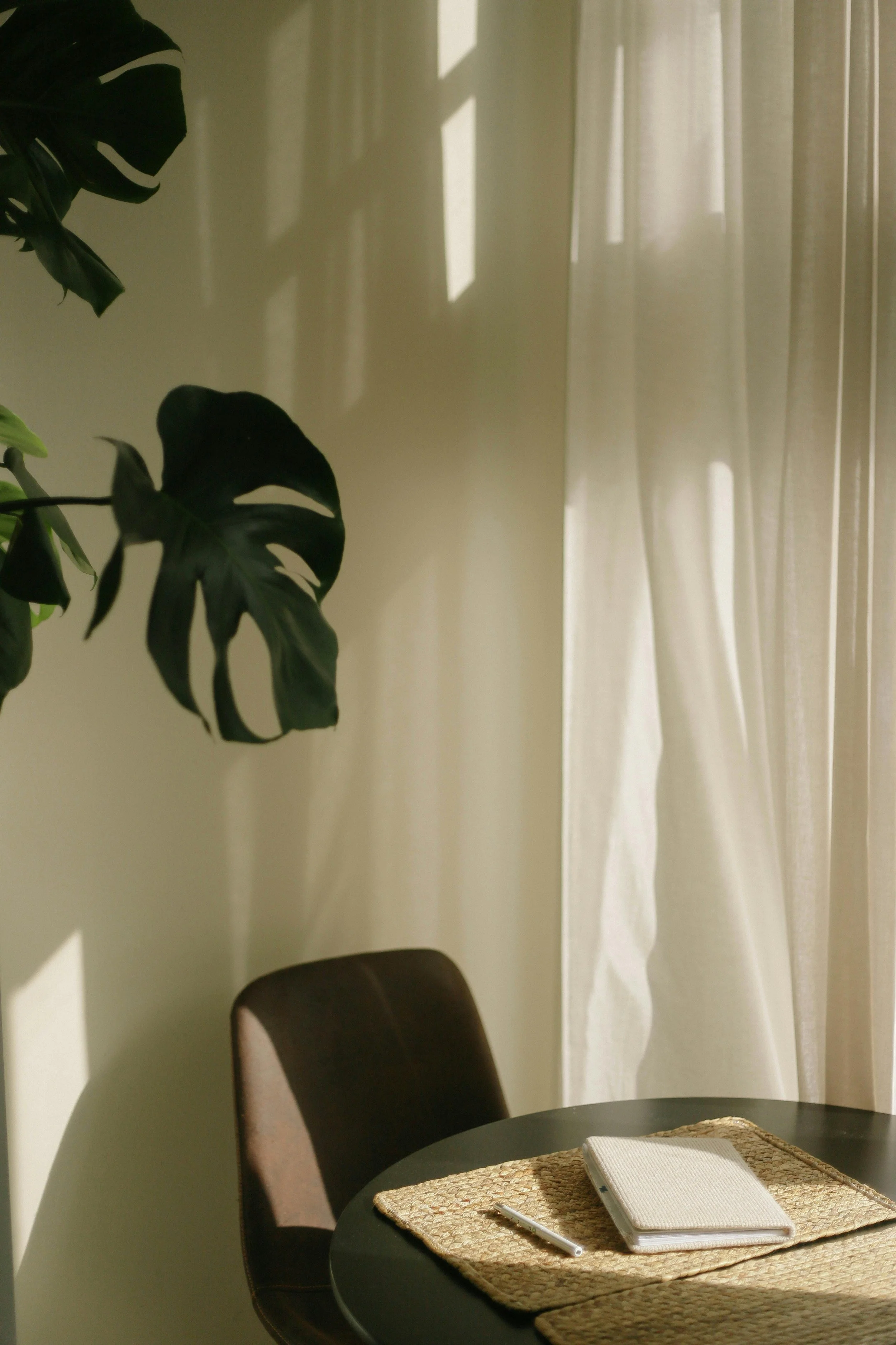 A dining room corner with a black round table, a chair, a notebook, and a pen on a woven placemat. Sunlight filters through curtains, creating shadows on the wall and curtains.