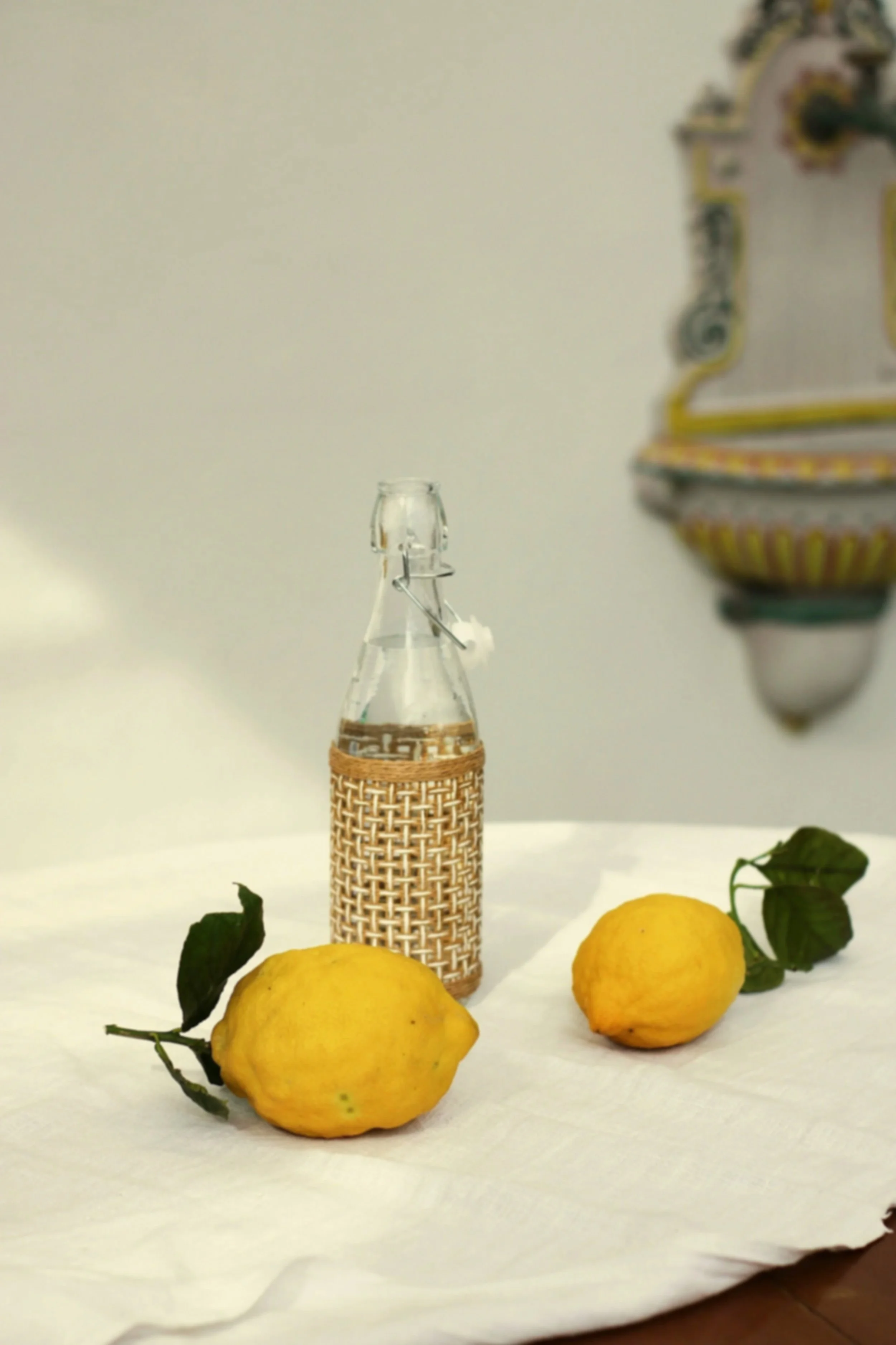 Three yellow lemons with black leaves on a white surface, with a small glass bottle and an ornate decorative wall hanging in the background.
