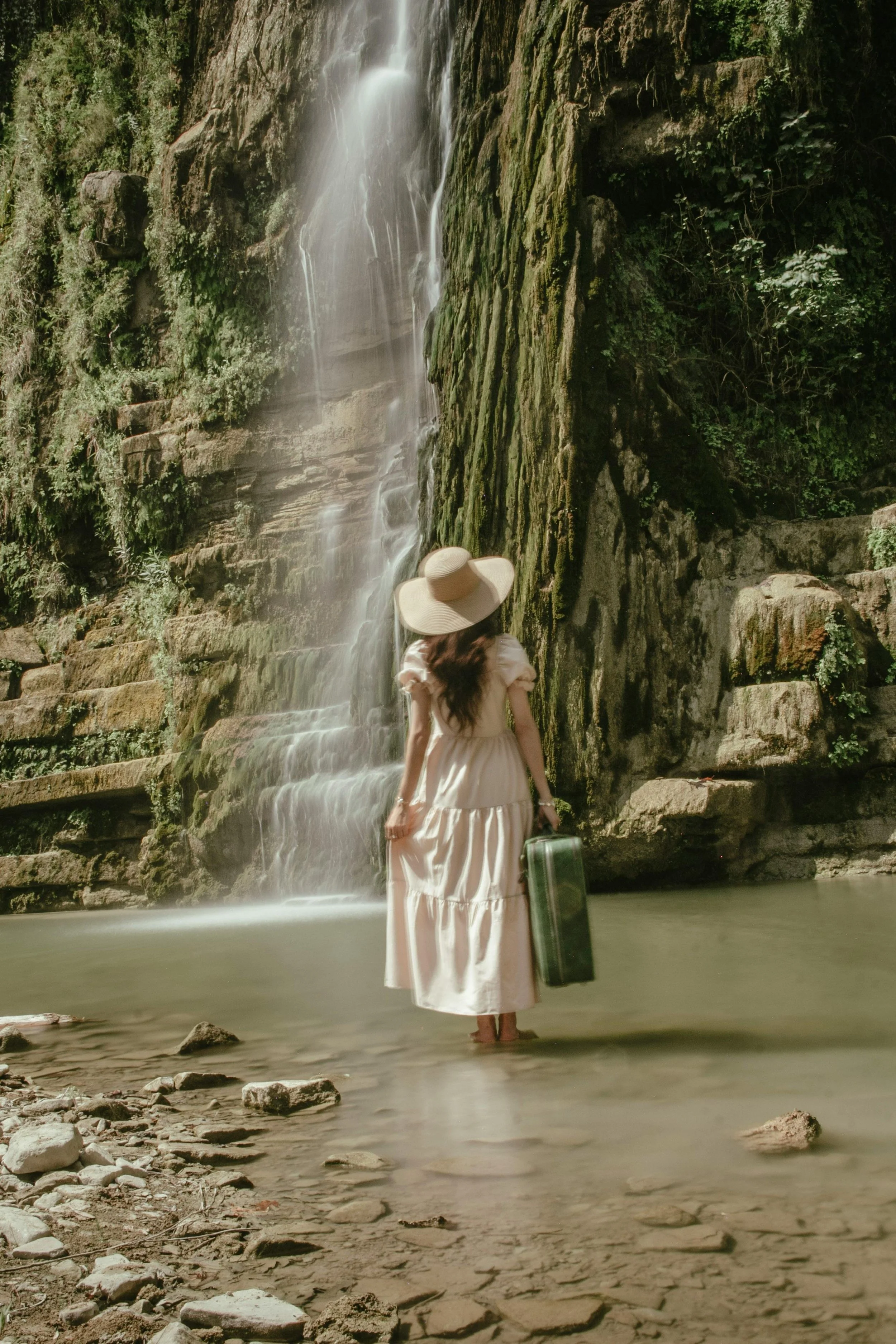 Woman standing in a river with a waterfall behind her, wearing a long white dress and a wide-brimmed hat, holding a green suitcase.