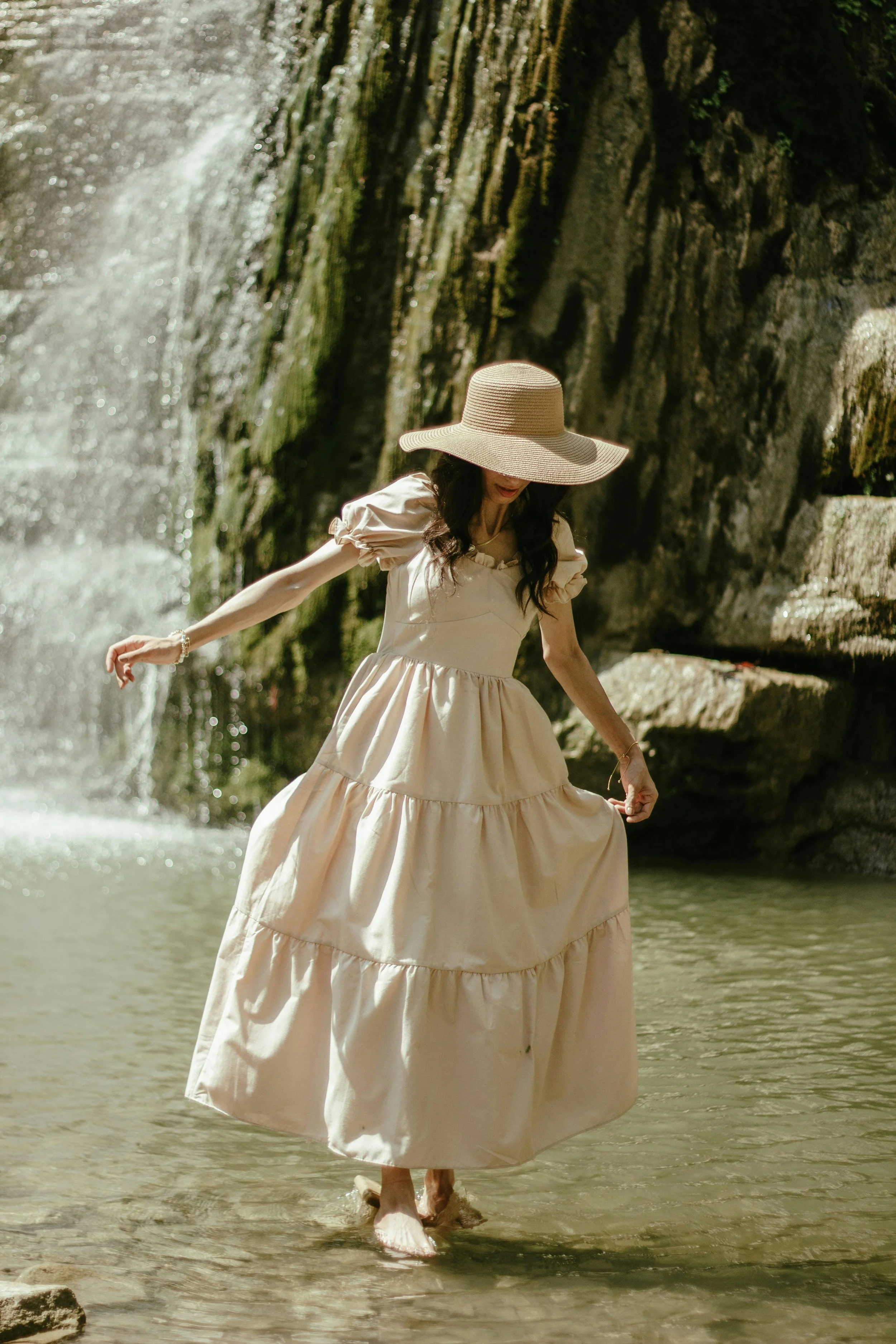 A woman wearing a wide-brimmed straw hat and a long, flowing cream-colored dress is walking in shallow water near a waterfall.
