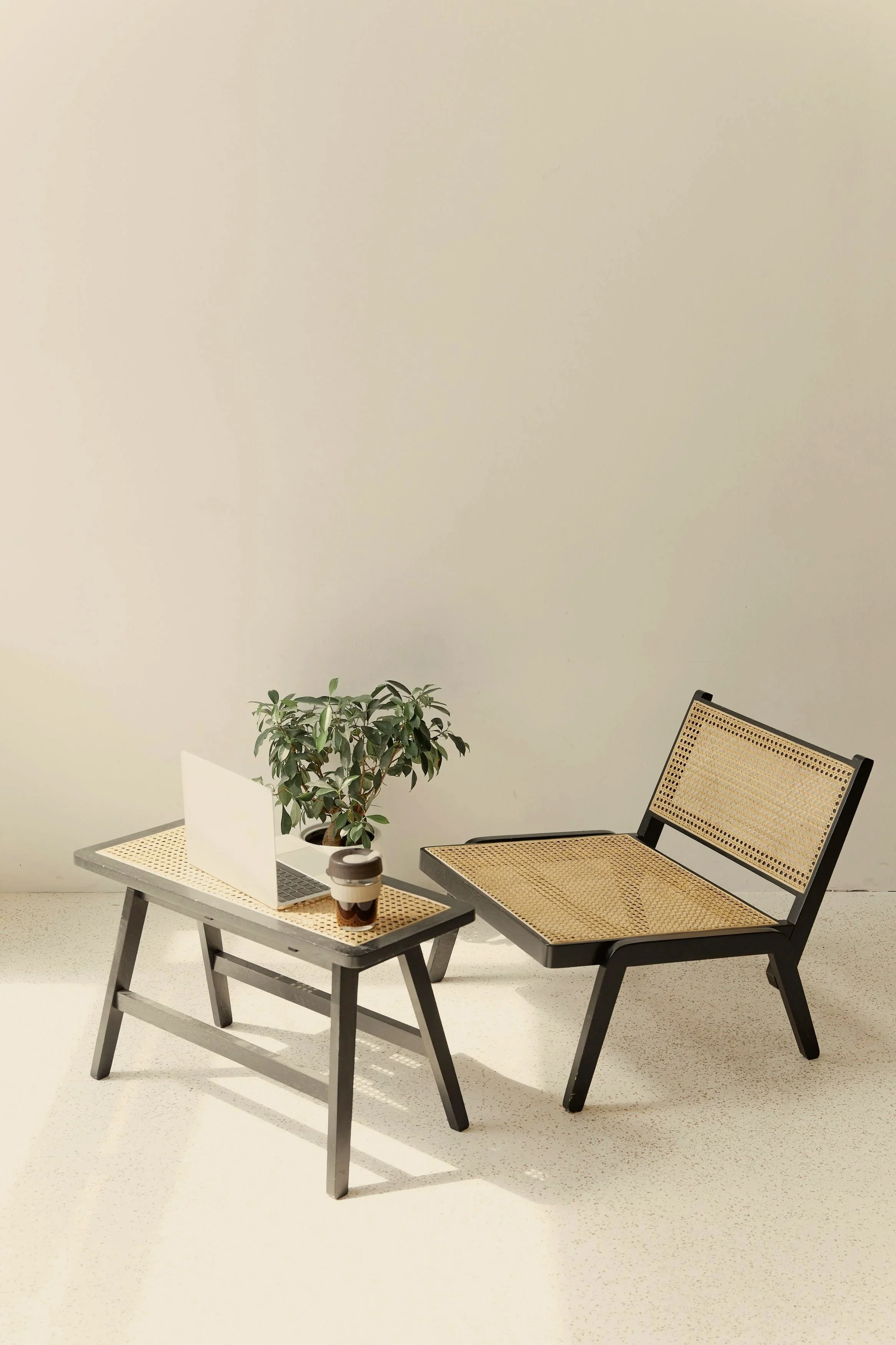 A minimalist desk arrangement with a laptop, a coffee cup, and a potted plant on a small table, next to a chair with a woven seat and backrest, all set against a plain, light-colored wall.