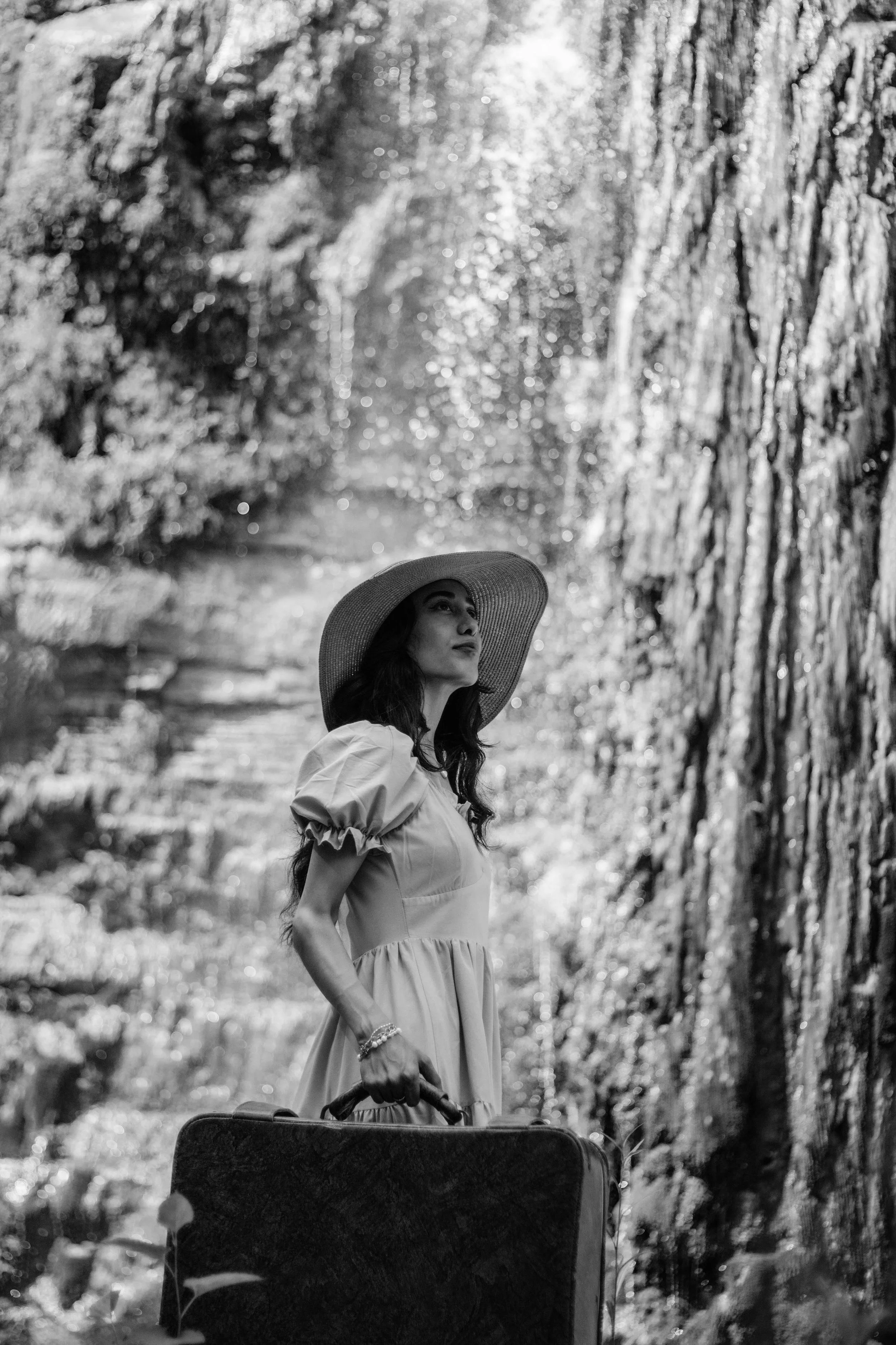 A woman in a wide-brimmed hat, wearing a light-colored dress with puffed sleeves, standing outdoors on a sunny day in a wooded area, holding a suitcase.