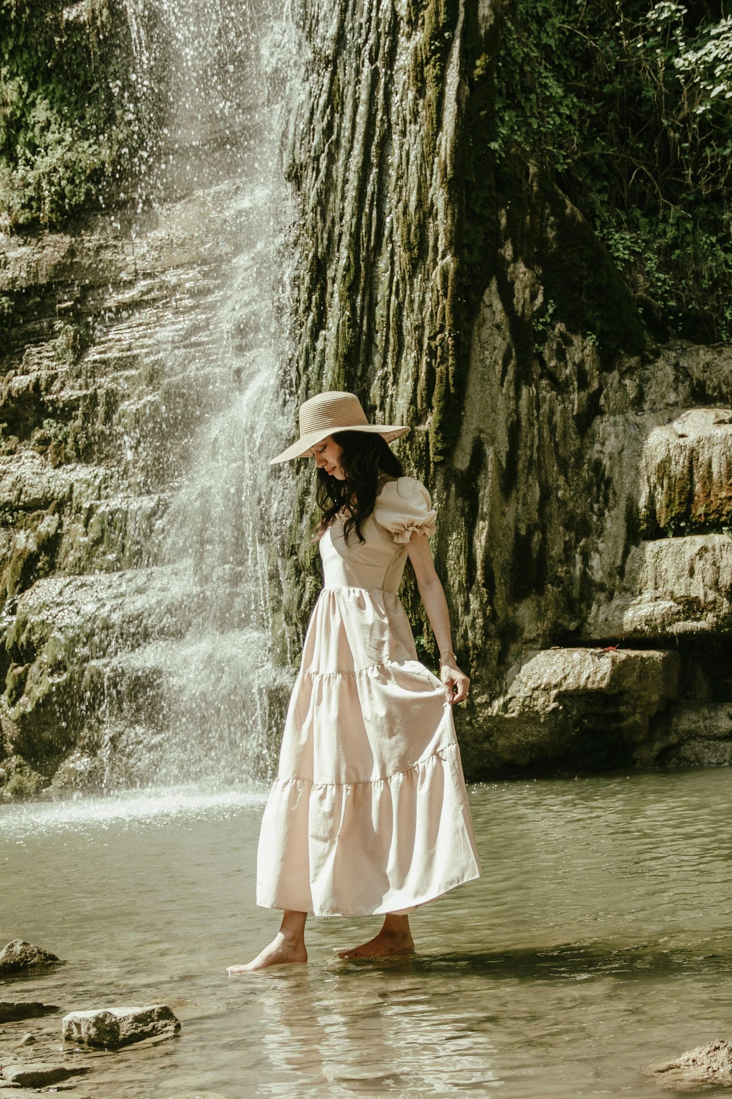 A woman in a beige dress and wide-brimmed hat standing in shallow water near a waterfall.