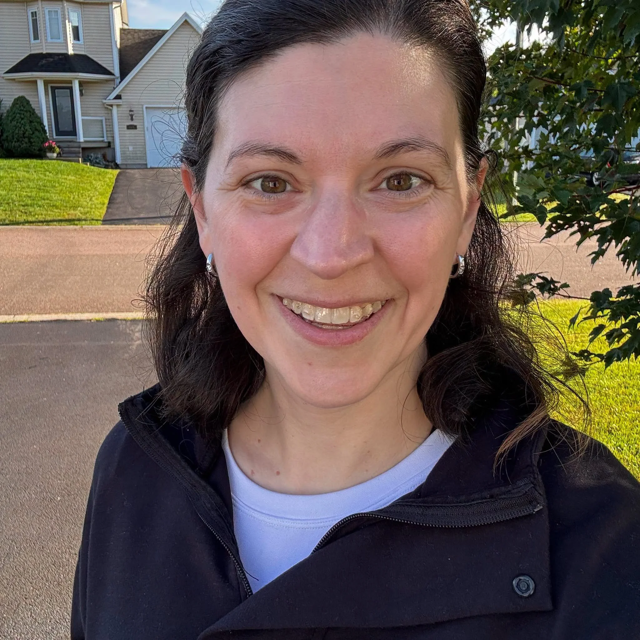 A woman with dark hair smiling outdoors in front of a house and trees.