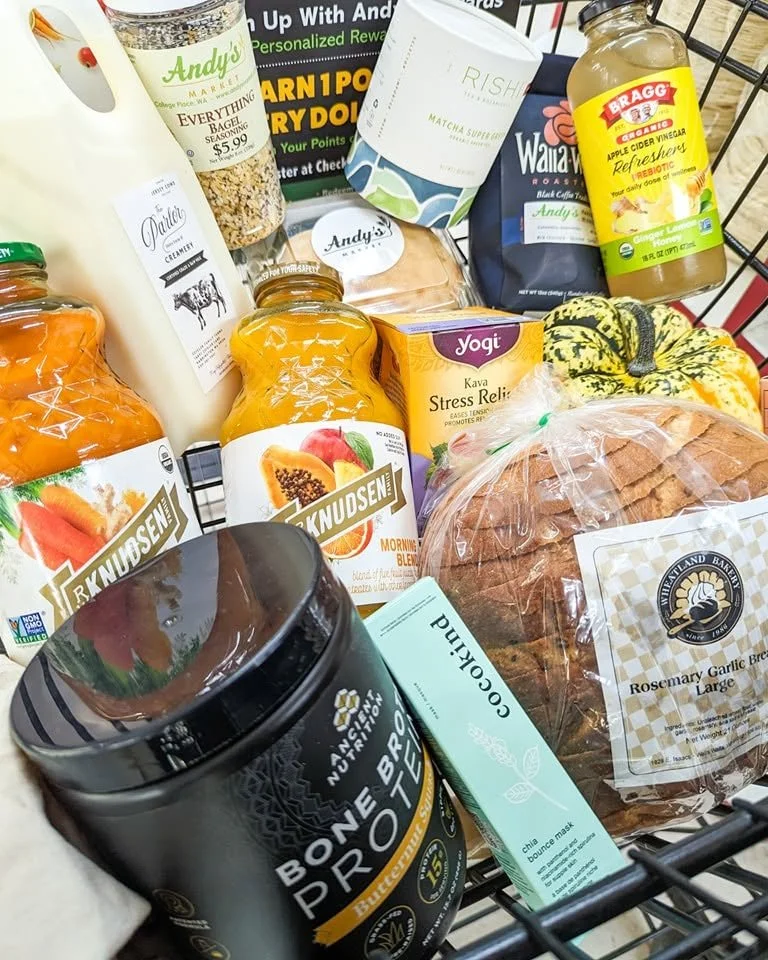 A grocery cart filled with various items including bread, juice bottles, a pumpkin, a mask, and other packaged foods and beverages at Andy's Market.