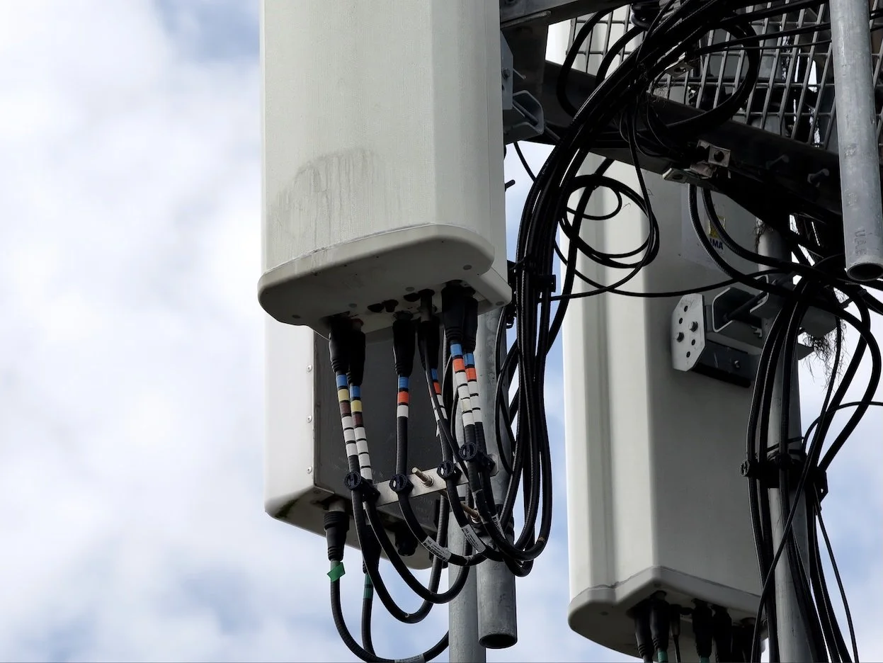 Close-up of outdoor telecommunications equipment, including white electrical boxes and multiple black cables connected to it, mounted on a metal pole against a cloudy sky background.