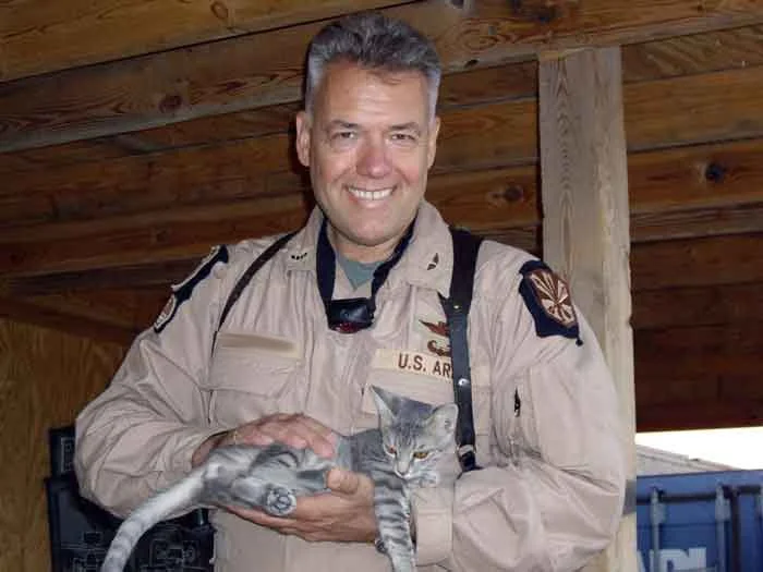 A man in a U.S. Army uniform smiling and holding a gray tabby cat in a wooden outdoor shelter or porch.