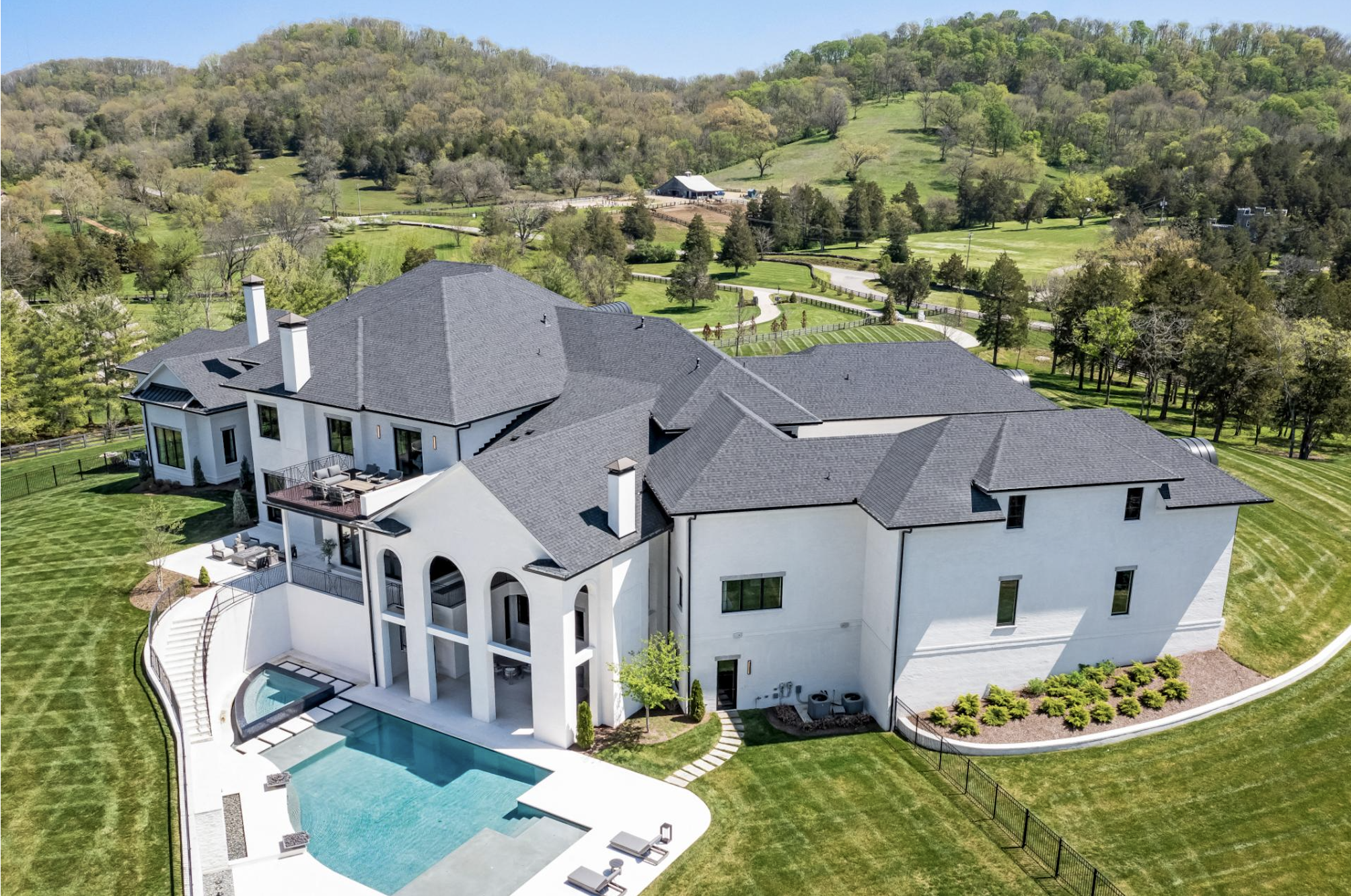 A large white mansion with multiple levels, balconies, and black chimneys, featuring a backyard with a swimming pool and poolside seating, surrounded by a well-maintained lawn and lush greenery, with rolling hills and trees in the background under a clear blue sky.