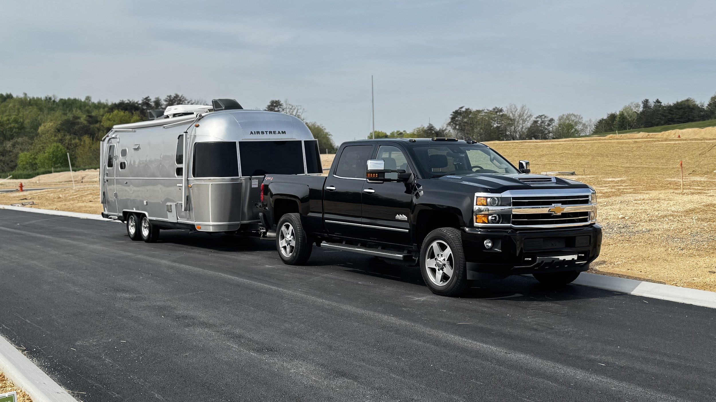A black Chevrolet truck towing a silver Airstream trailer on a paved road during daytime, with a background of sparse trees and a partly cloudy sky.