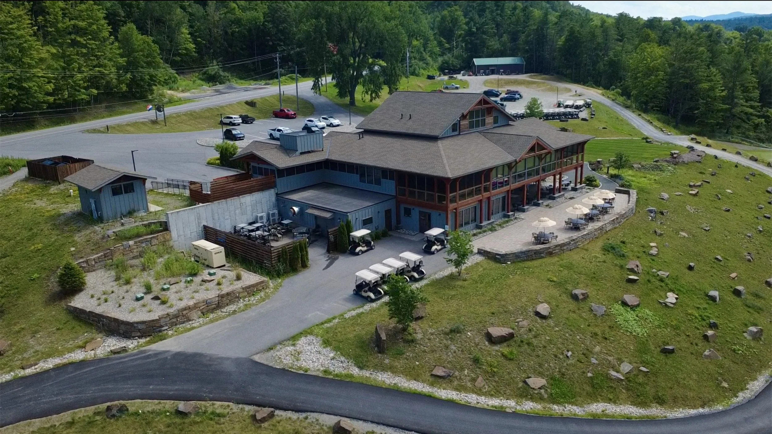 An aerial view of a large building with a wooden exterior, surrounded by a parking lot with several golf carts and cars. There is an outdoor patio area with tables and umbrellas, and a landscaped garden with rocks and plants. A forested area and a road are visible in the background.