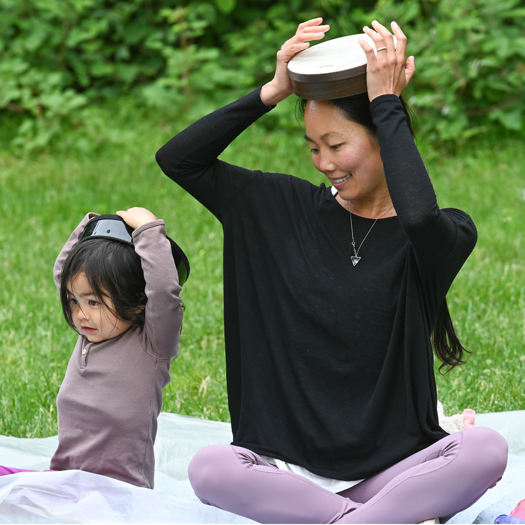 A woman and a young girl sit on a picnic blanket outdoors in a green park. The woman is smiling and placing a wooden drum on her head, while the girl is also holding a similar drum above her head.