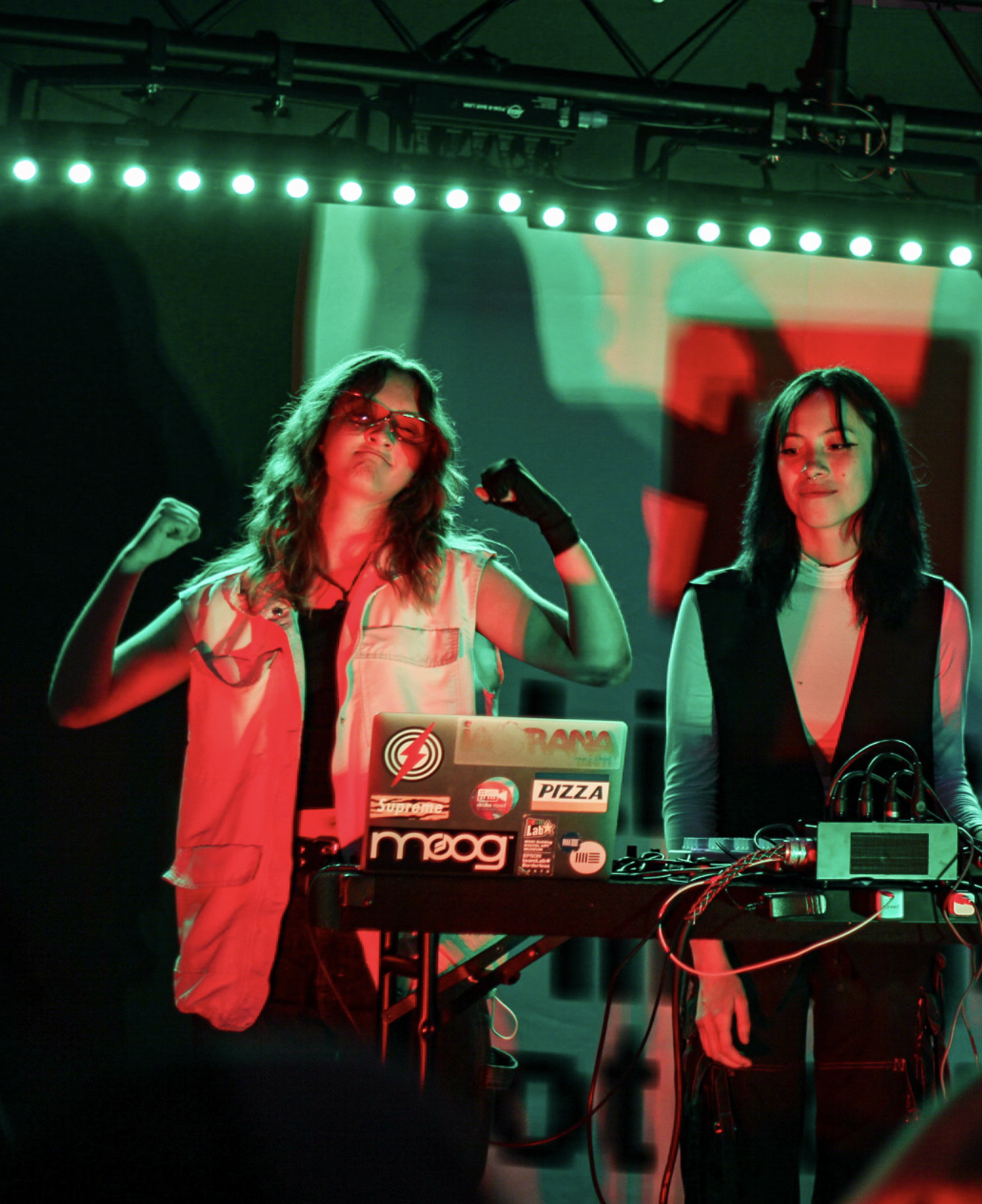 Two women are DJing at a music event, with one flexing her arms and the other standing beside her, surrounded by DJ equipment and colorful lighting.