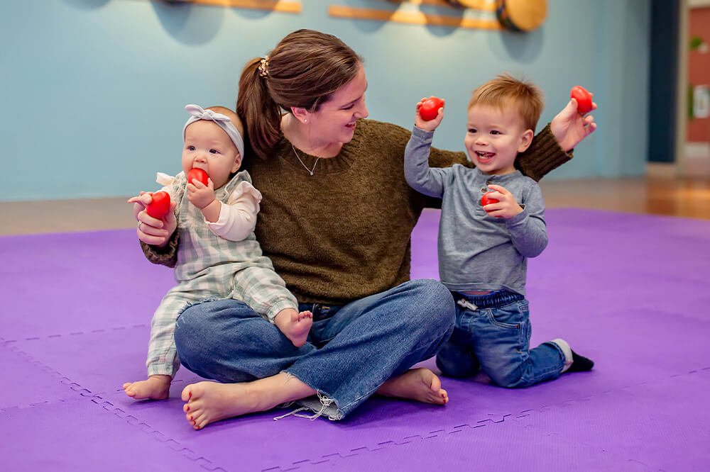A woman sitting cross-legged on a purple mat with a young girl on her lap and a young boy standing beside her, all holding red balls and smiling during playtime indoors.