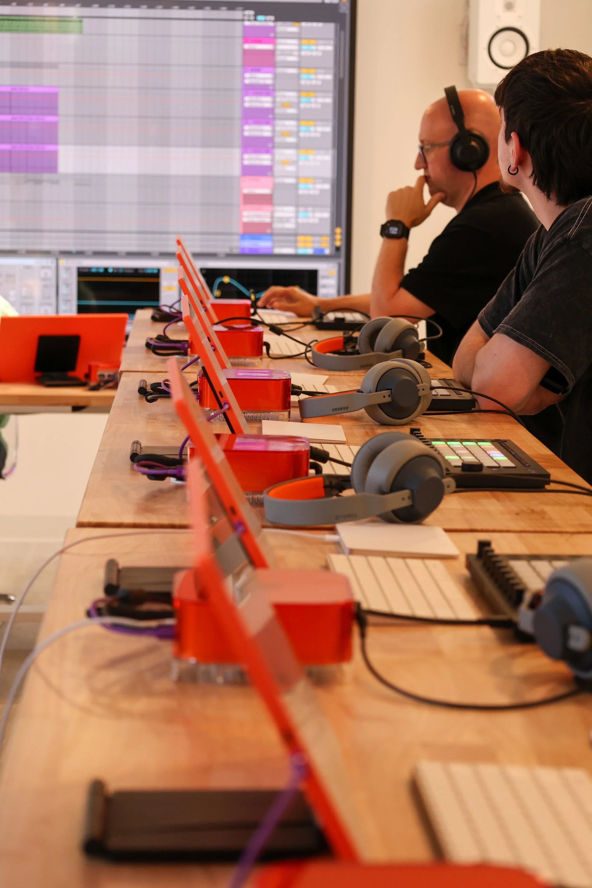 Two people working at a music production setup with a large screen displaying digital audio workstations, headphones, and electronic musical equipment on a wooden table.