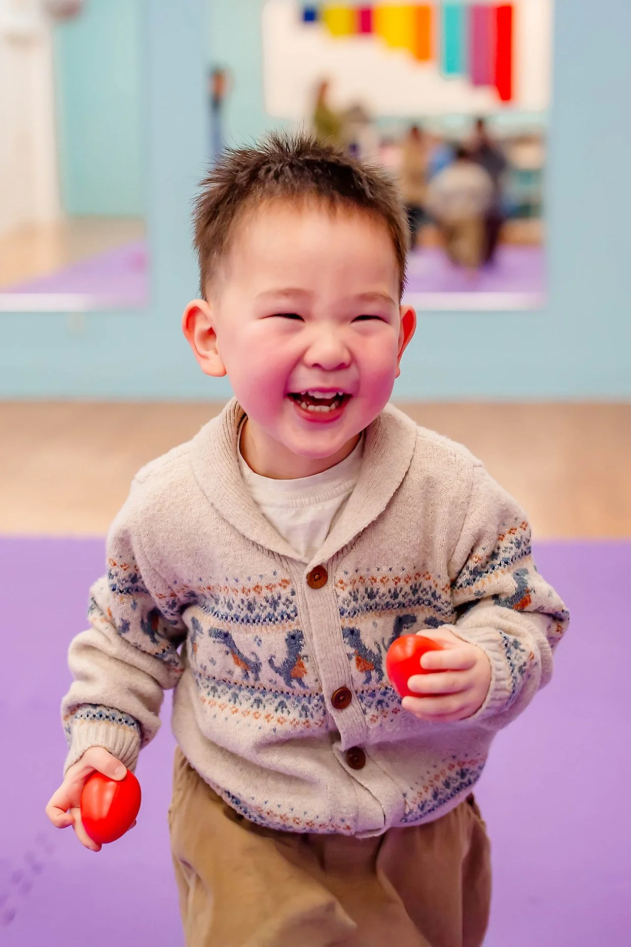 A young boy with short brown hair, laughing and holding red balls in both hands, standing in a colorful indoor play area with purple flooring and blurred background of other children and adults.